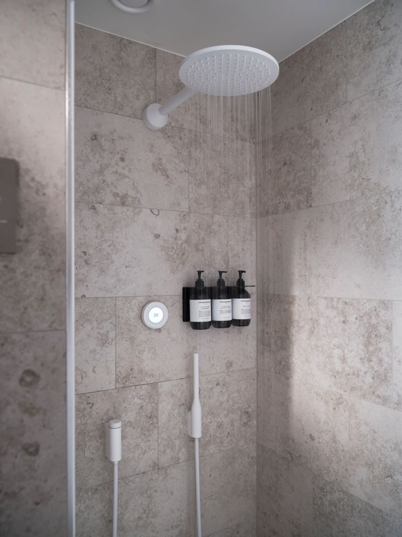 Water streams from a modern rain shower head onto light stone tiles, beside a digital temperature control, wall-mounted toiletries, and two white handheld shower wands.