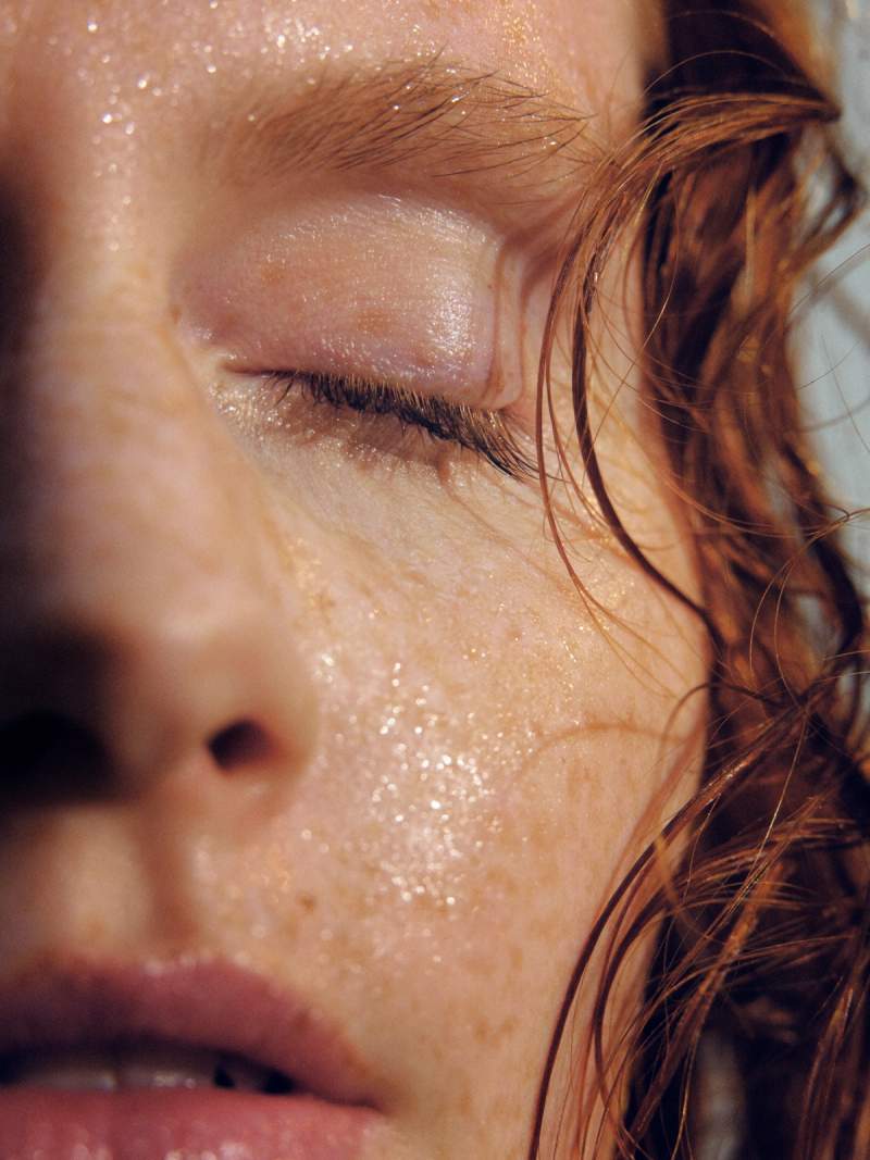 Close-up of a person's freckled face, glistening with moisture, with a closed eye and wet reddish-brown hair.