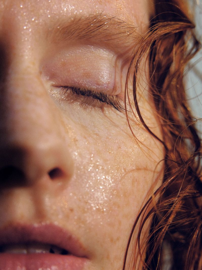 Close-up of a person's freckled face, glistening with moisture, with a closed eye and wet reddish-brown hair.