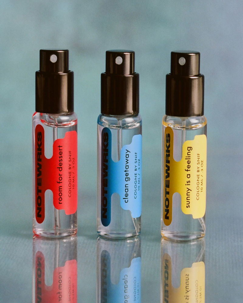 Three NoteWrks cologne spray bottles, labeled "room for dessert" (red), "clean getaway" (blue), and "sunny is a feeling" (yellow), stand on a reflective surface.