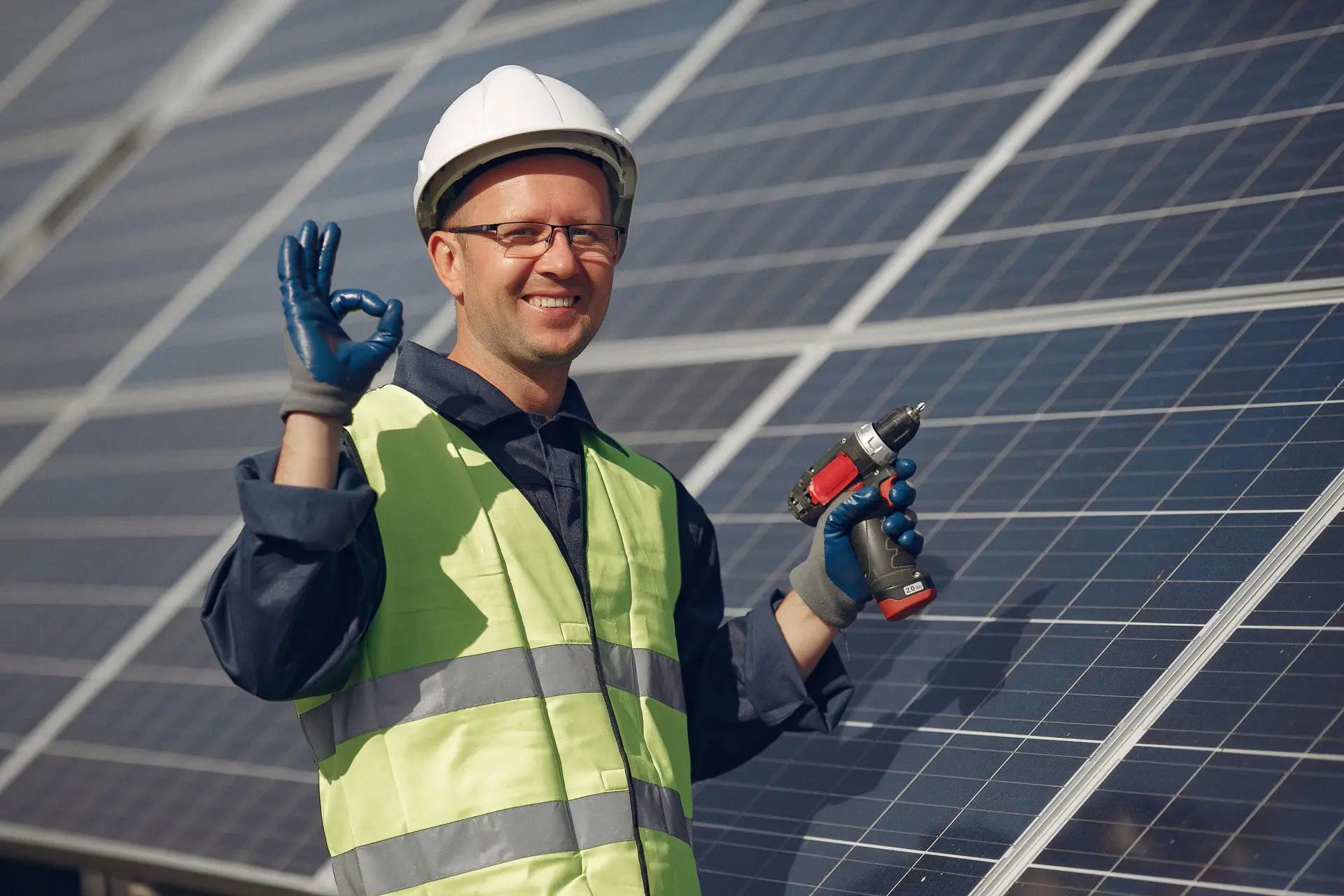 Solar installer in a hard hat and safety vest giving an OK sign in front of panels