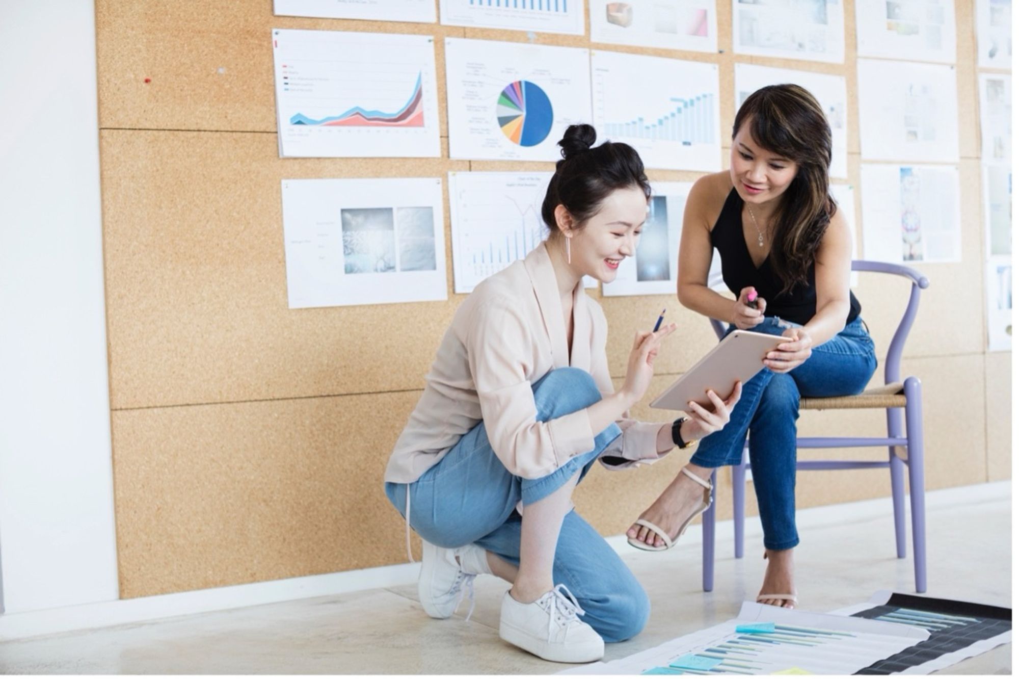 Two women looking at a tablet together in front of a wall covered in charts and graphs.