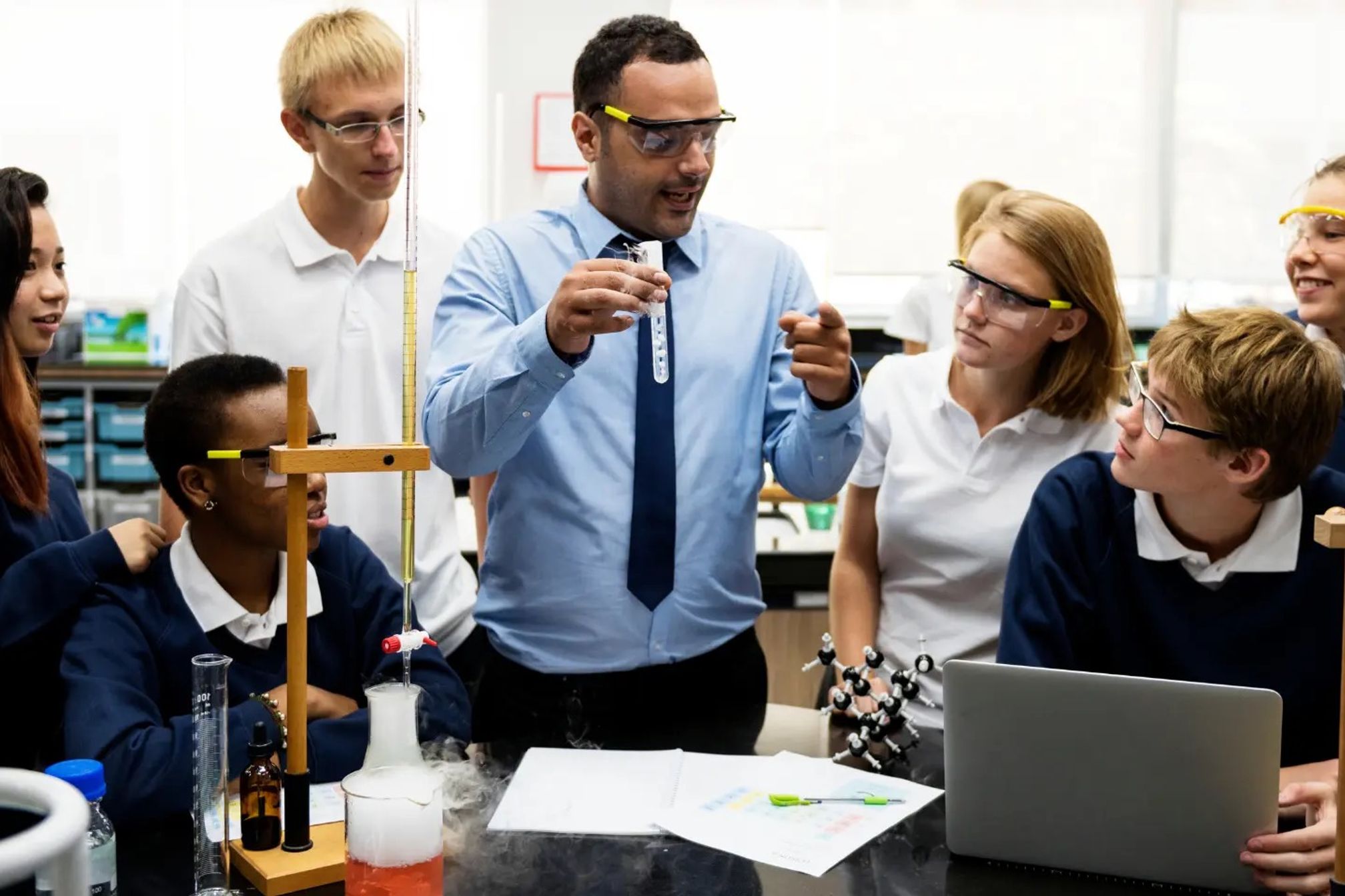 A teacher and his students in a classroom lab, they are wearing safety goggles and there is chemistry apparatus around them, he is holding a test tube and speaking to his students.