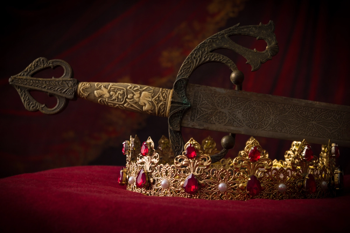 A gold decorative sword and a gold crown with red jewels and white pearls on a red velvet cushion.