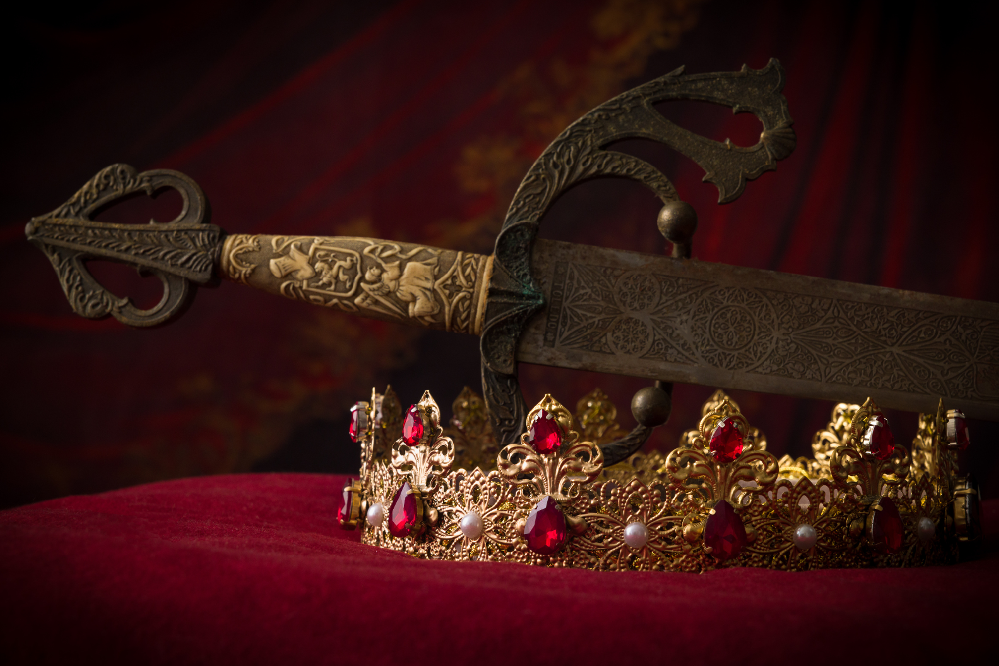 A gold decorative sword and a gold crown with red jewels and white pearls on a red velvet cushion.