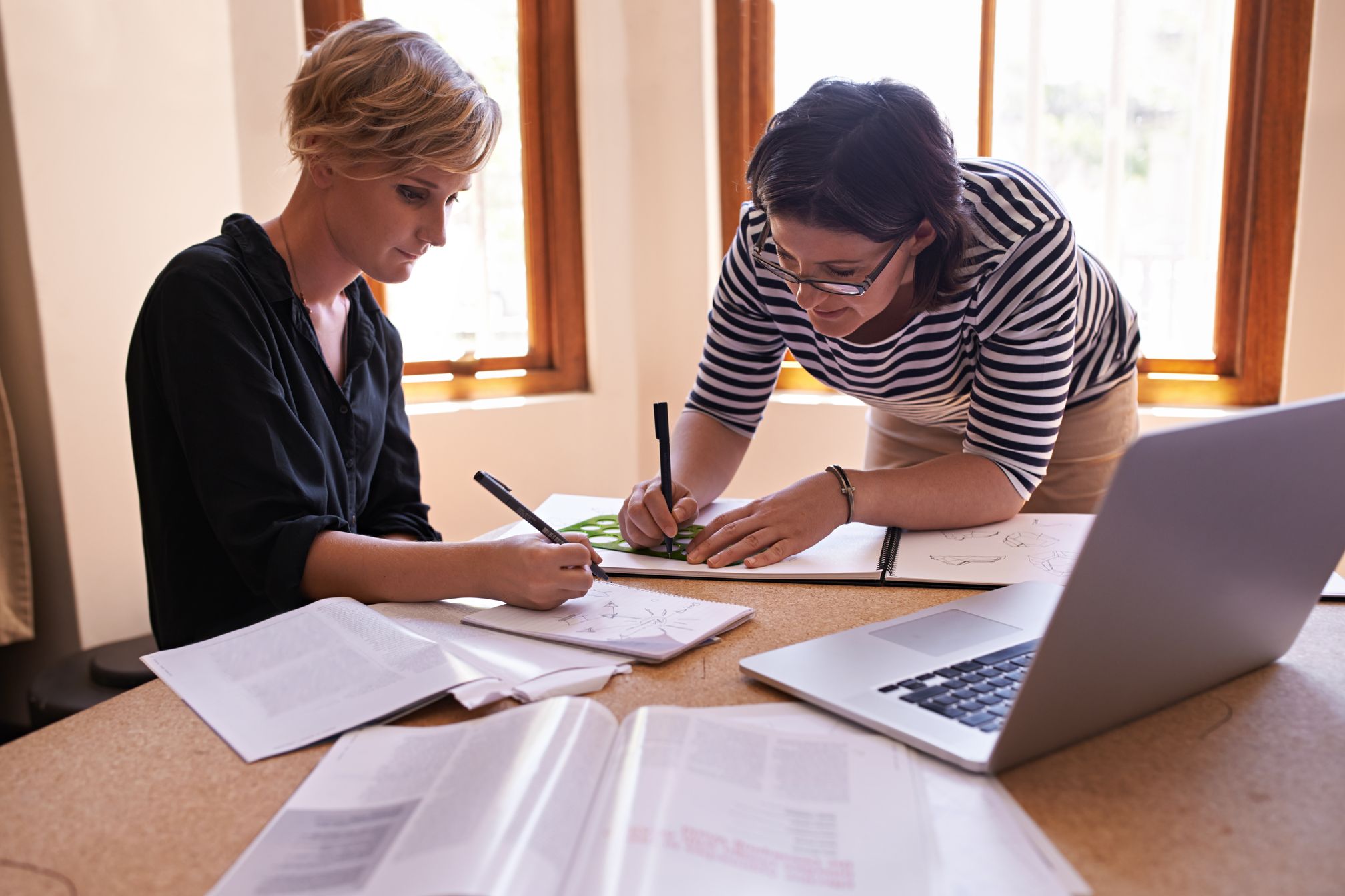Two women at a desk writing working together a laptop is on the desk.