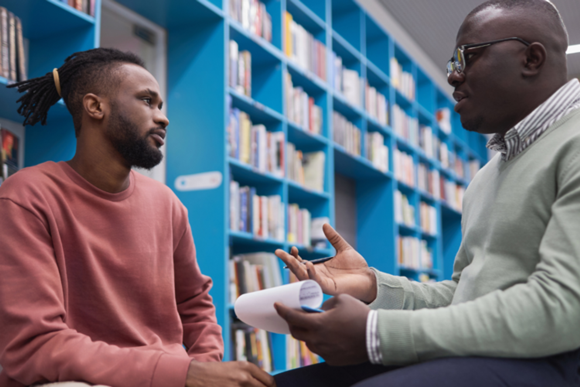 Two men are seated facing each other, one is speaking to the other holding a clipboard and a pen and is dressed smartly, the other is listening, he is dressed more casually. They are in a room full of books on bookcases.