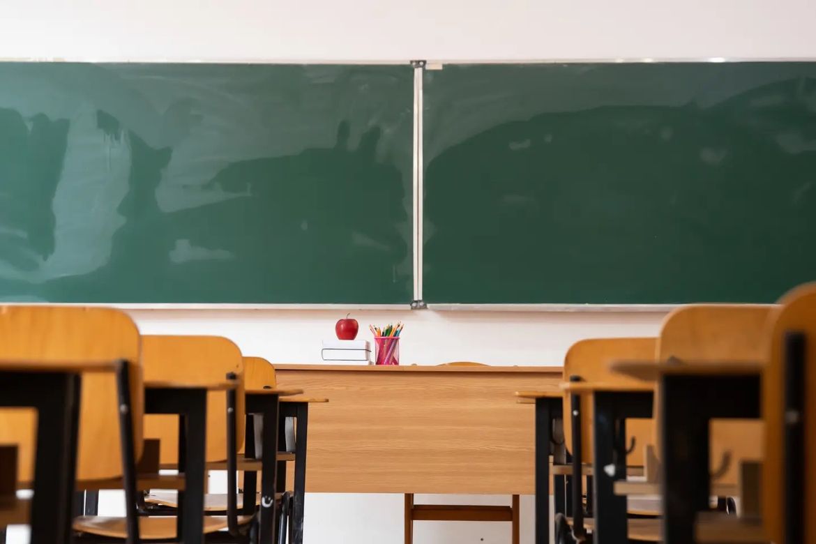 An empty classroom with wooden desks and chairs with two green chalkboards on the wall