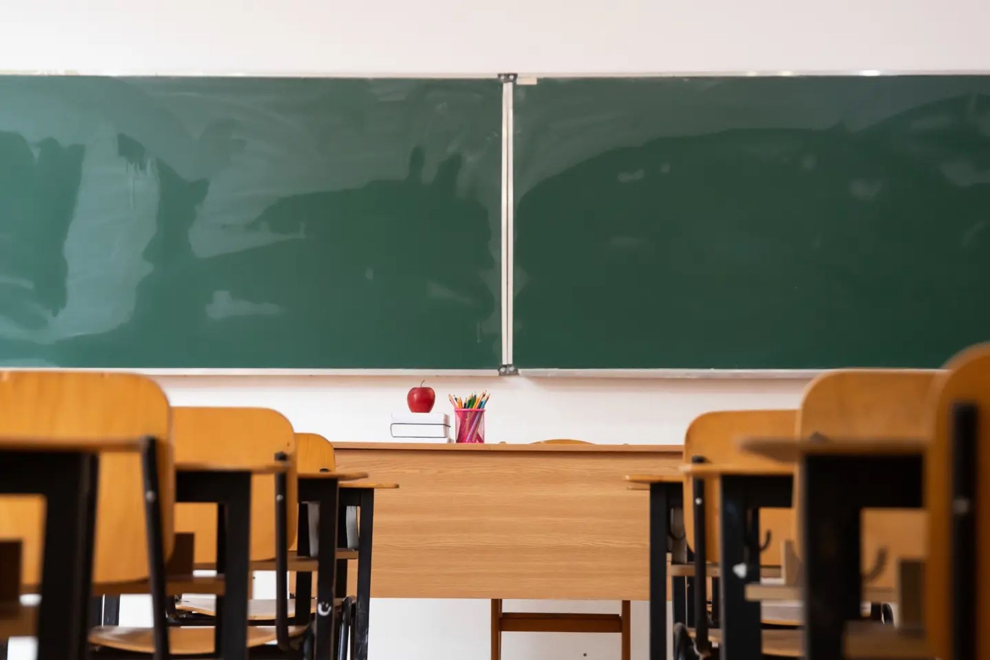 An empty classroom with wooden desks and chairs with two green chalkboards on the wall