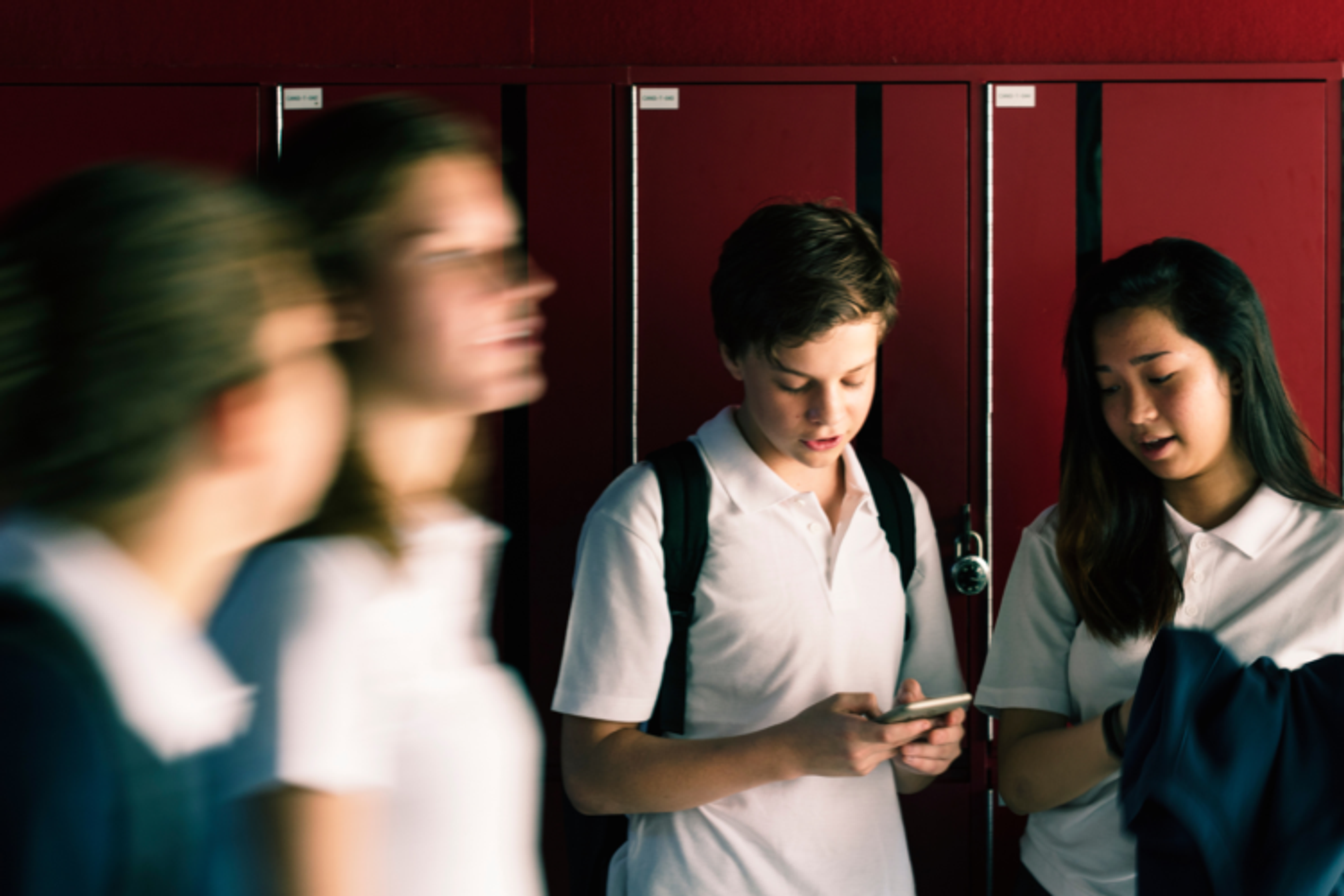 Teenage school children in a corridor with lockers along the walls, looking at a phone together.