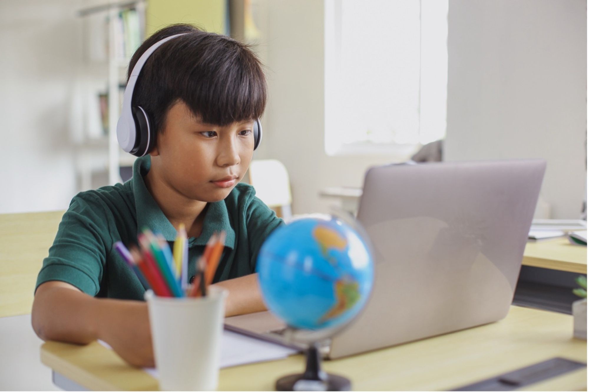 A young boy wearing headphones sitting at a table looking at a laptop screen there is a globe and a pot of pencils beside him.