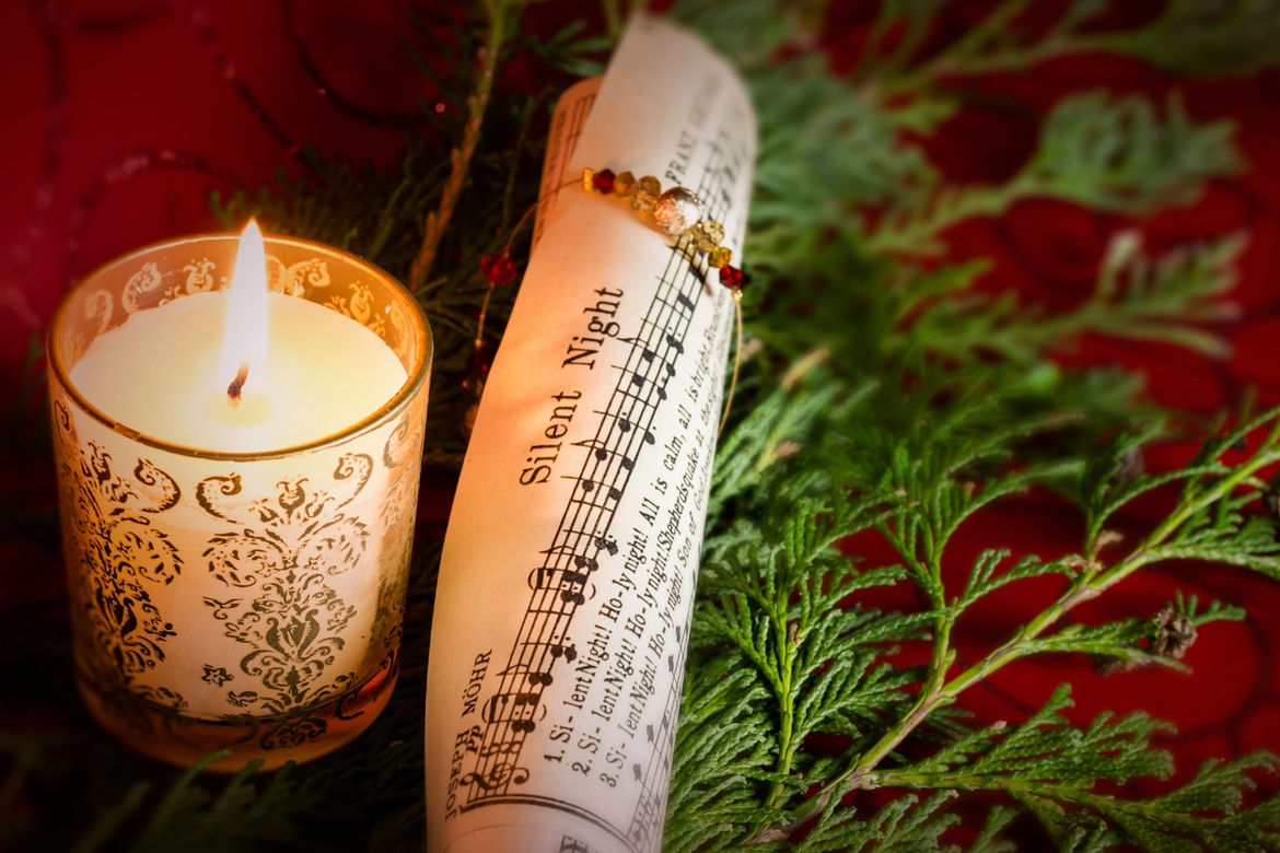 A lit candle in a decorative jar beside a curled up music score for "Silent Night" placed on a bed of green leylandii on a red table