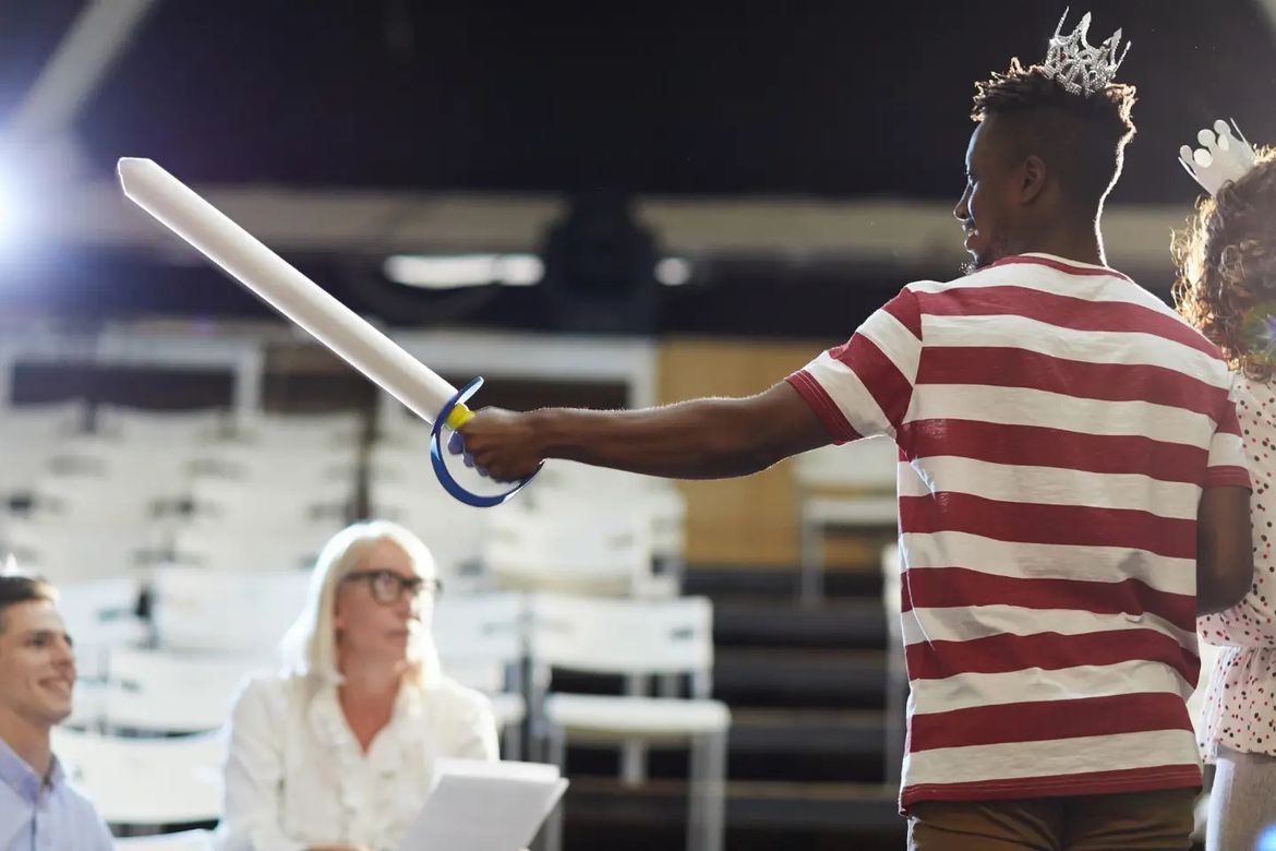 Students rehearsing on a stage with a sword prop and prop-style crowns. Watched by teachers