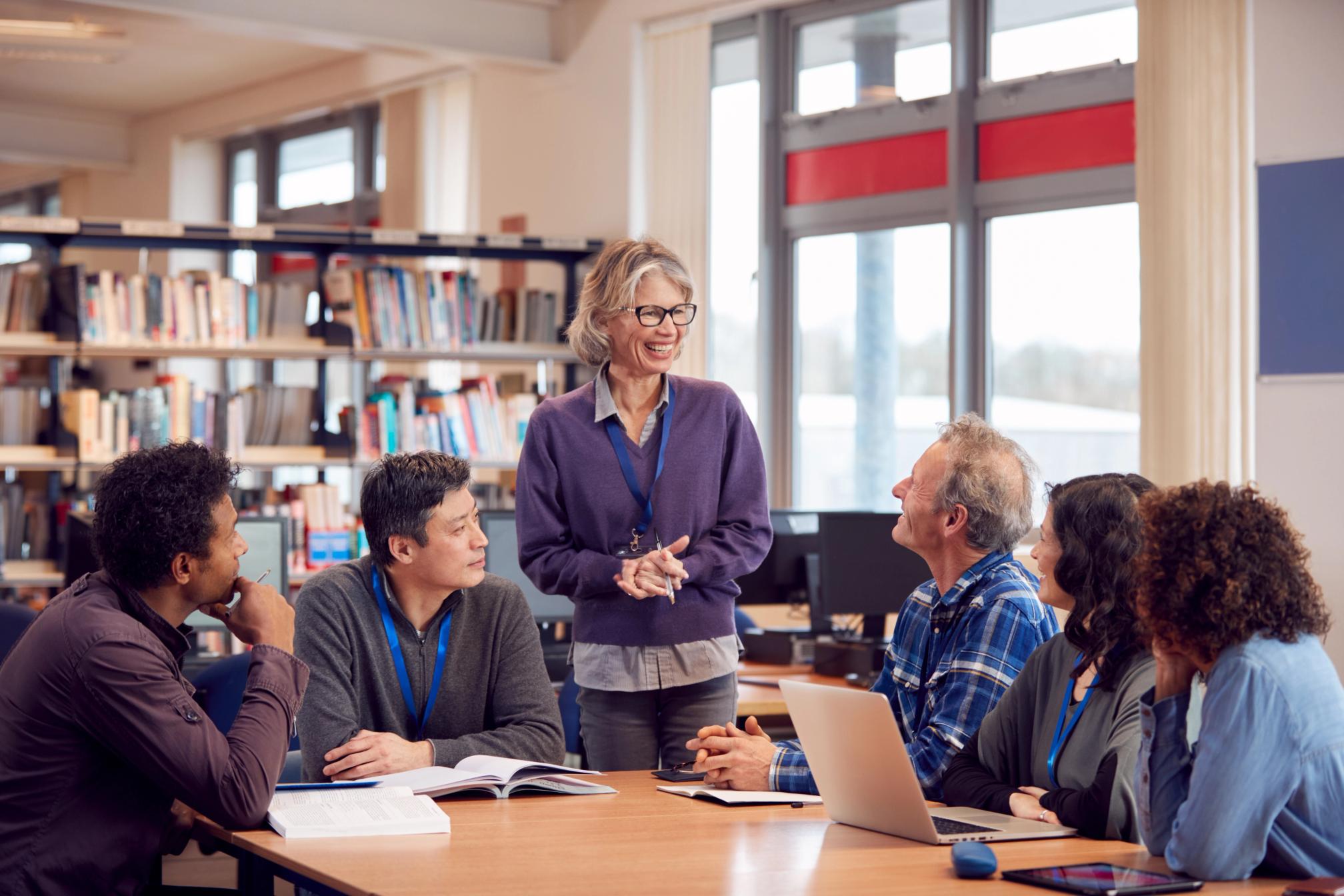 Adults sitting around a desk working together and talking in a library, one is standing