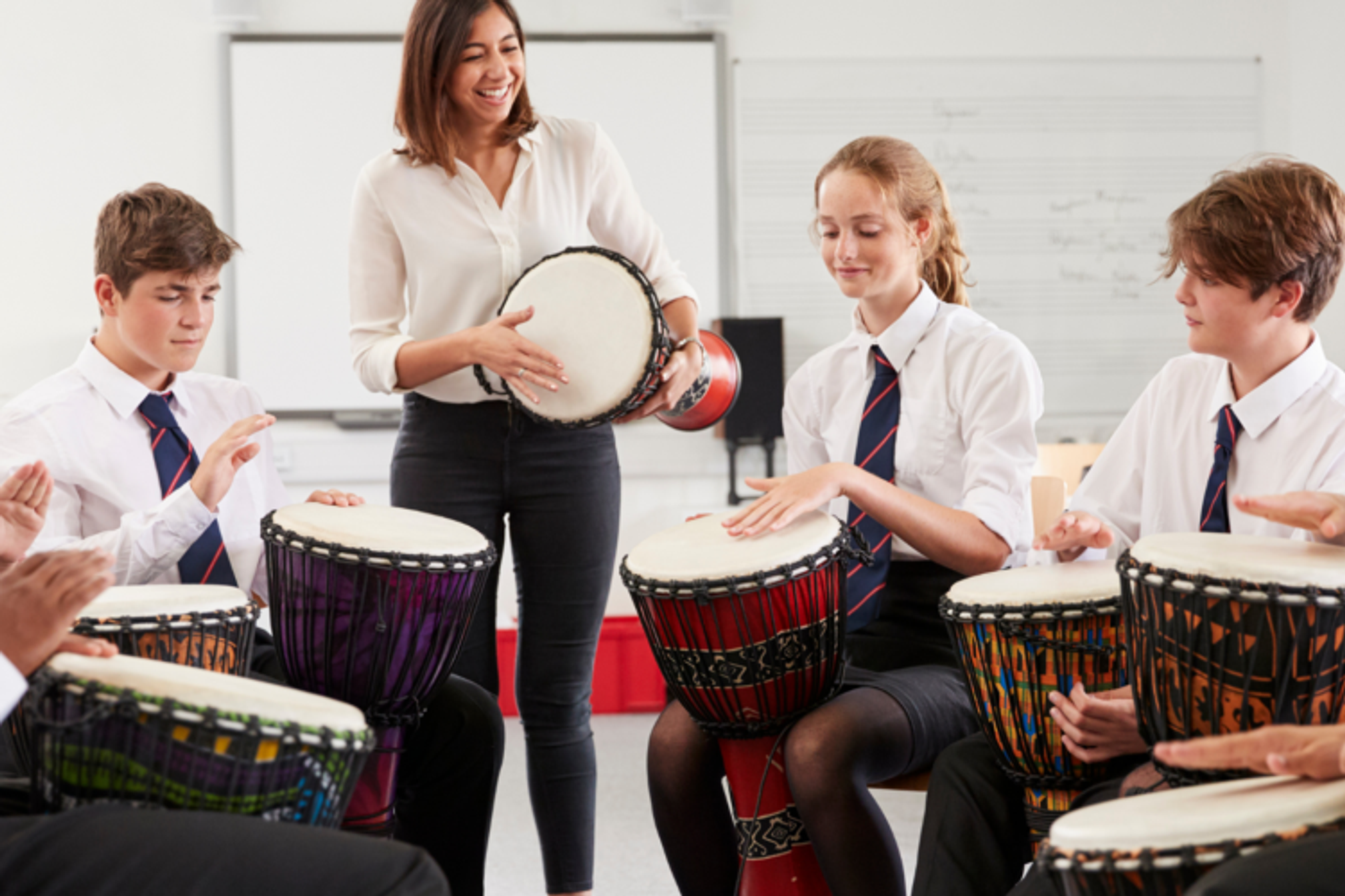 Secondary school children sitting in a group playing Djembe drums with their hands, they are in a classroom and their teacher is joining in whilst standing up