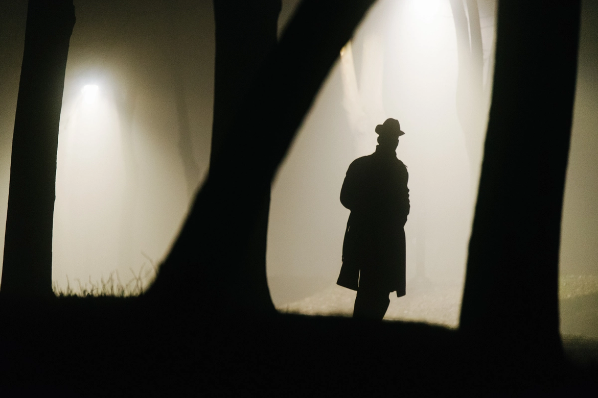A silhouette of a man in a brimmed hat wearing a long coat in some woods at night.