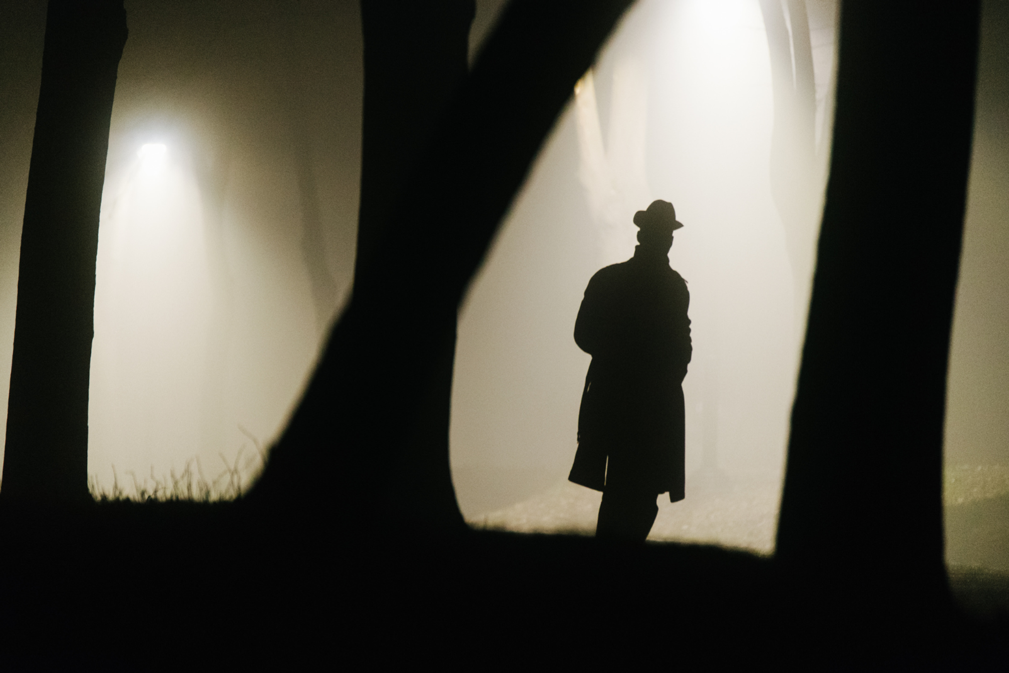A silhouette of a man in a brimmed hat wearing a long coat in some woods at night.