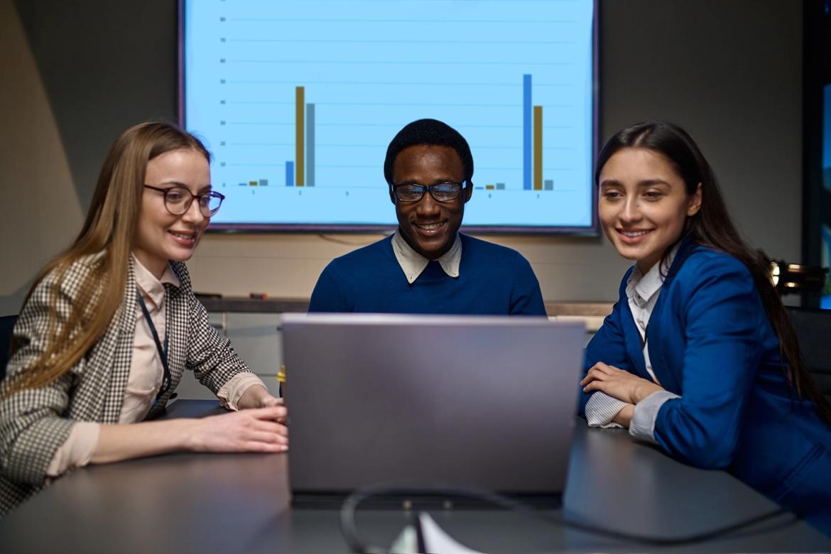 Three business people sitting around a laptop with a screen showing a bar chart behind them