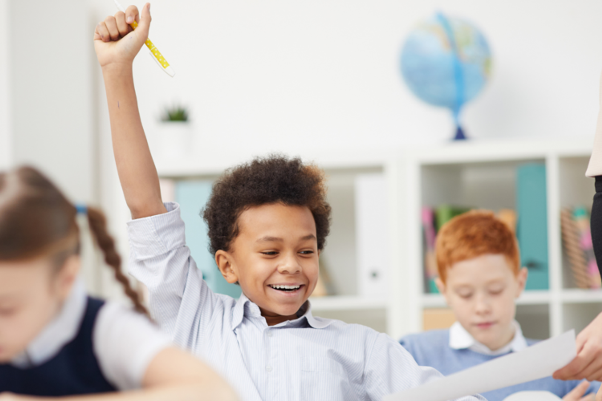A school boy in a classroom holding his hand up, a teacher is taking a sheet of paper from him.