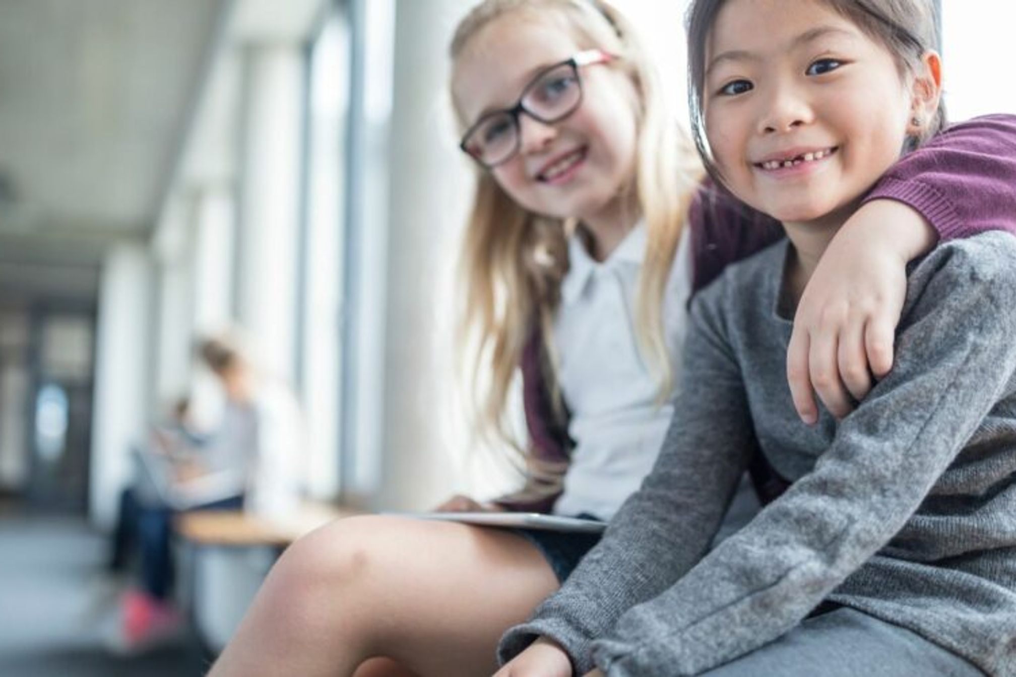 A young white schoolgirl sitting with her arm around a young asian schoolgirl in a school corridor looking at the camera smiling