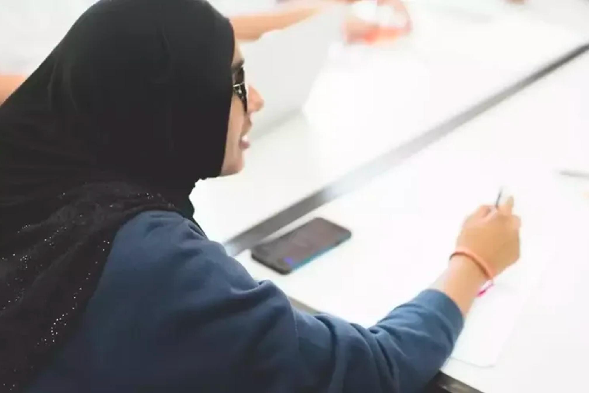 A schoolgirl wearing a hijab sitting at a table writing and speaking to someone across the table.