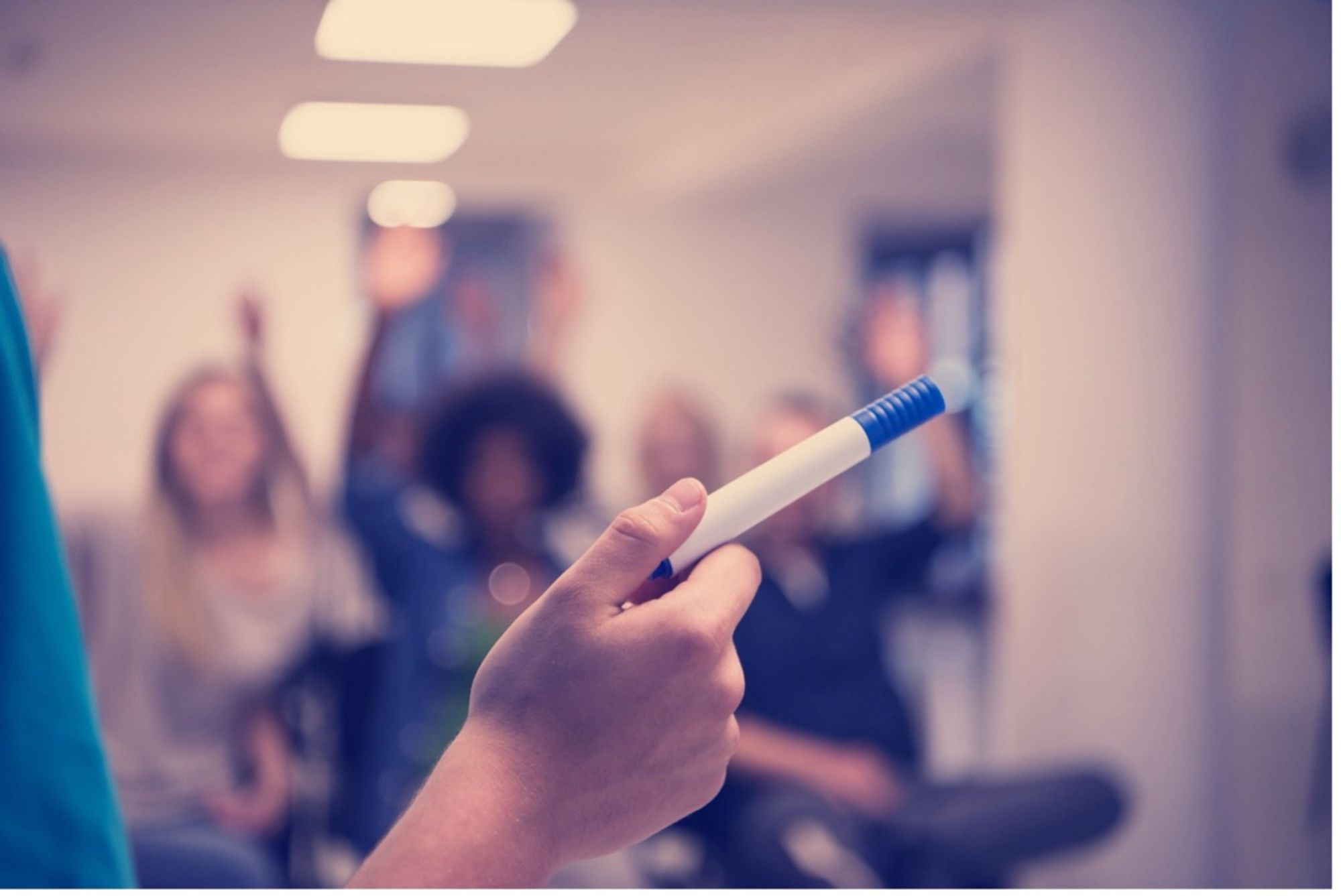 A hand holding a pen, possibly a teacher in front of a class, students raising hands.