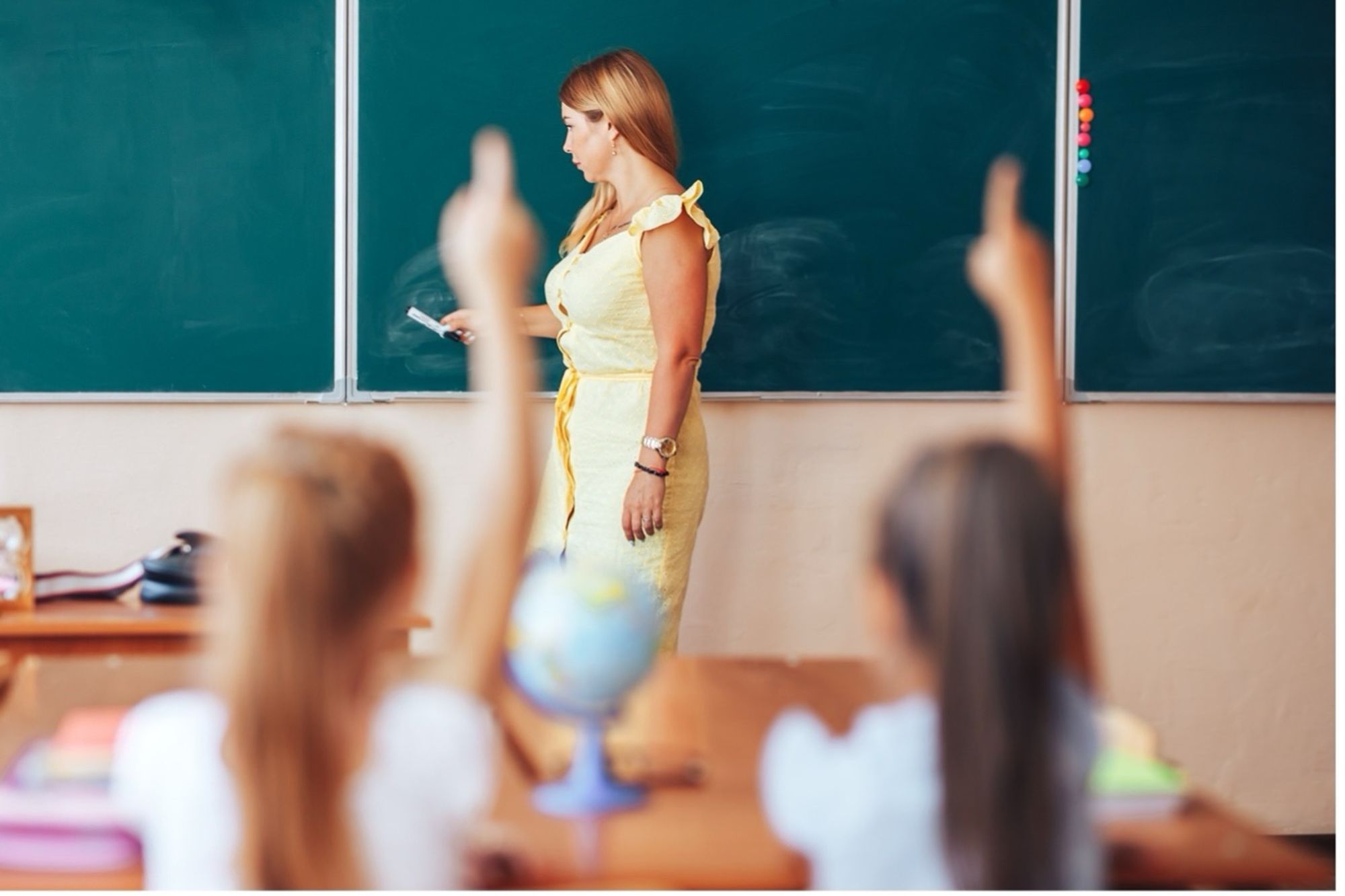 A teacher writing on a green chalkboard in a classroom students raising their hands.
