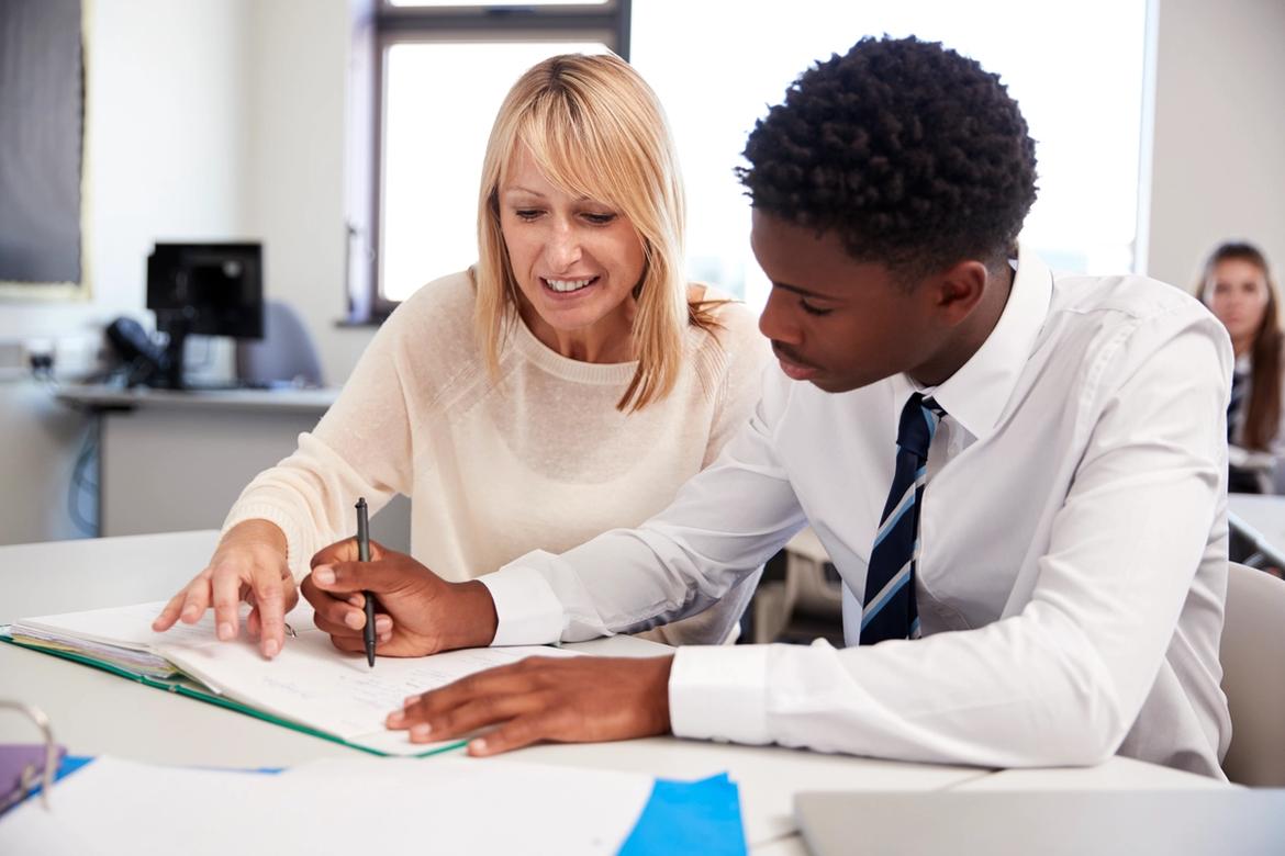 Teacher sitting at a desk in a classroom helping a teenage student.