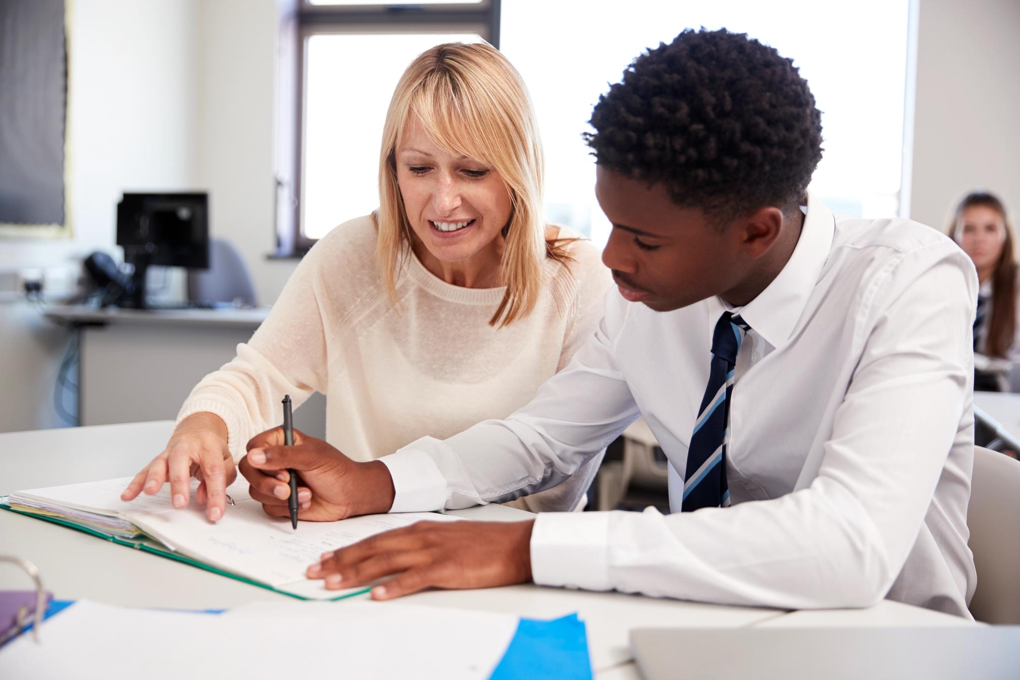Teacher sitting at a desk in a classroom helping a teenage student.
