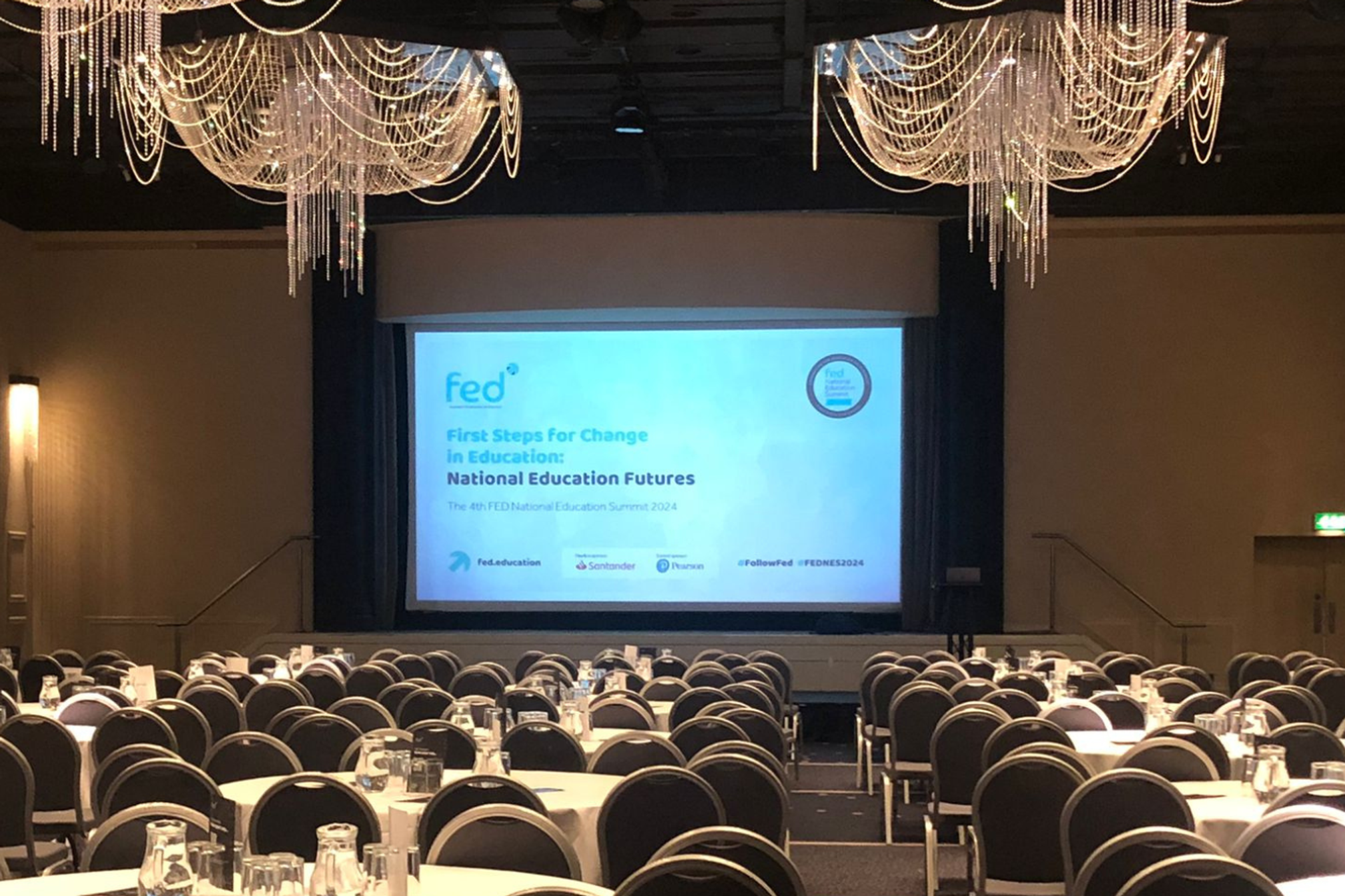 A conference hall with a presentation screen showing a title page for the "FED: First Steps for change in education, national education futures".