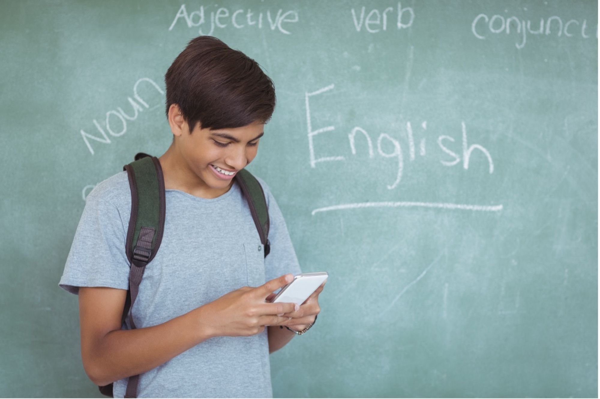 A boy standing in front of a green chalkboard with the words "English, noun, verb, adjective" written on the board, looking at his phone and smiling.