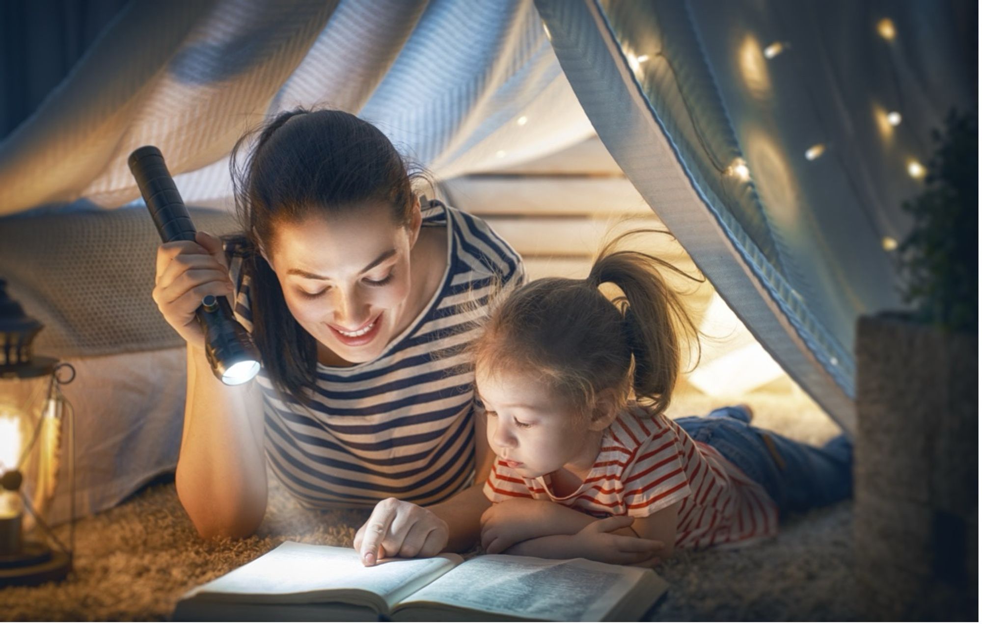 A young toddler girl with her young mother who is holding a flashlight reading a book together in a tent in the dark lit with lanterns and fairy lights..