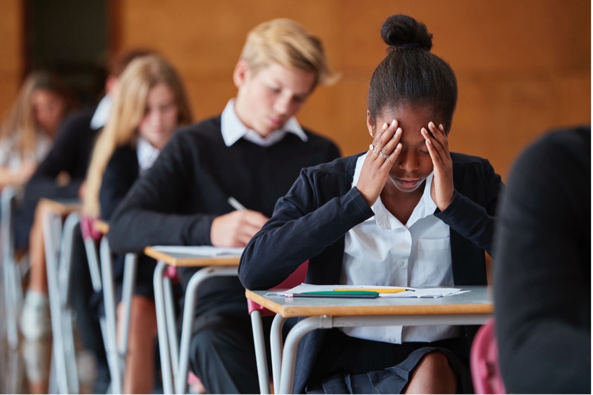 Teenage school children in uniform sitting in an exam hall, one student has her head in her hands.