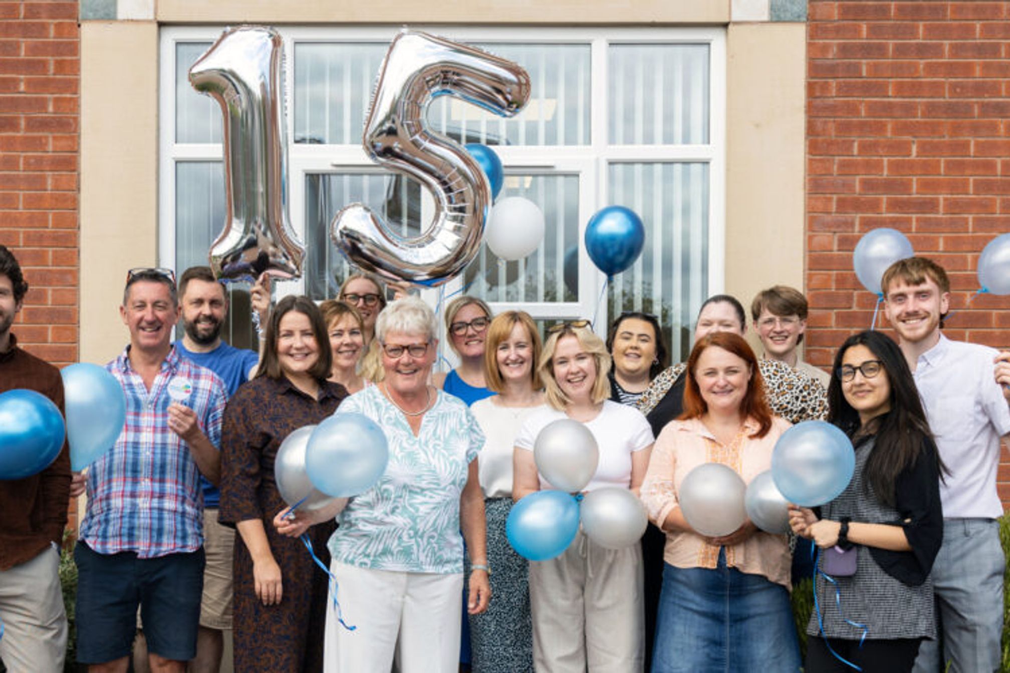 Group of people standing outside a building with balloons and a large balloon in the shape of a number 15