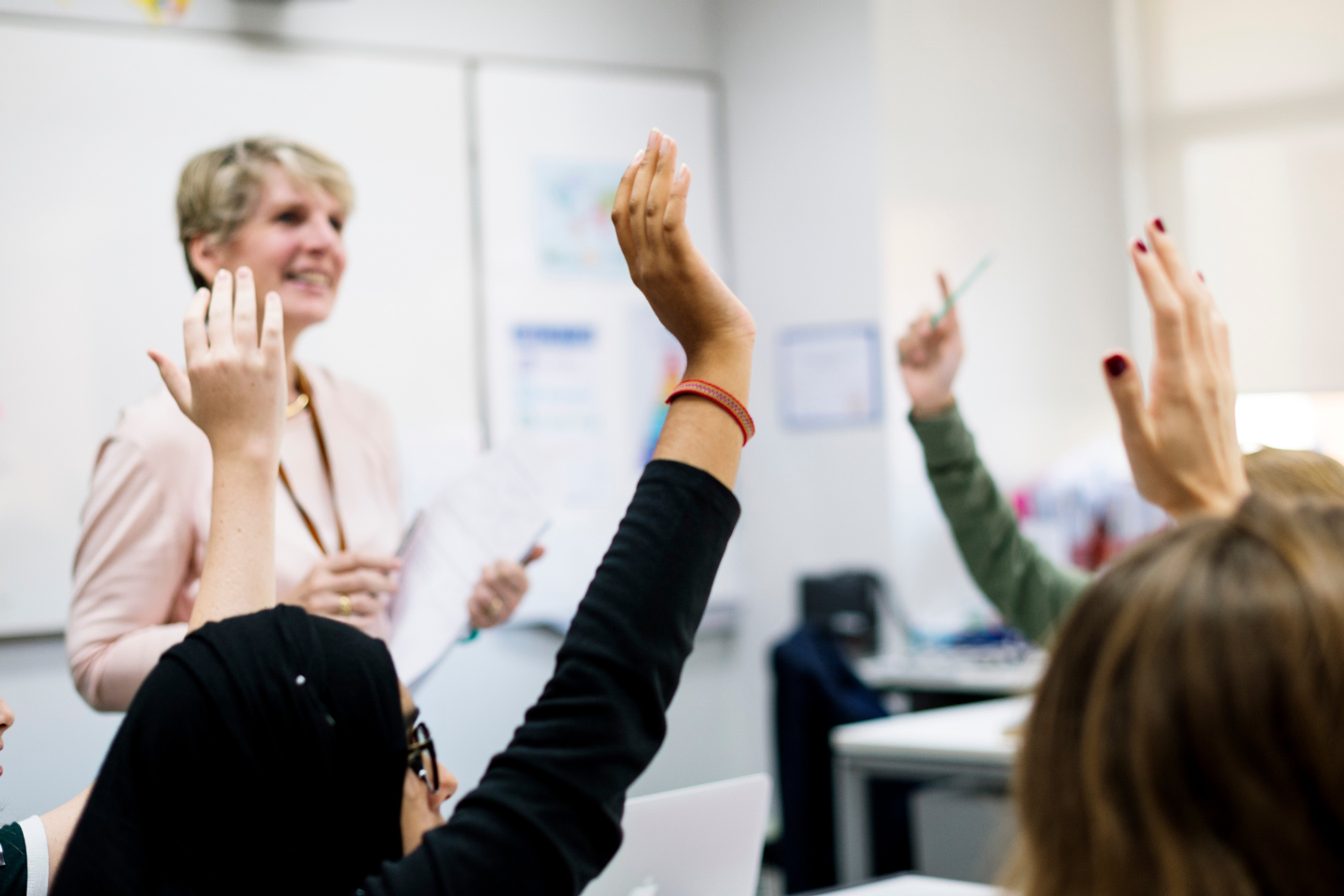 A group of secondary students in a classroom holding their hands up, a teacher is standing up in front of them..