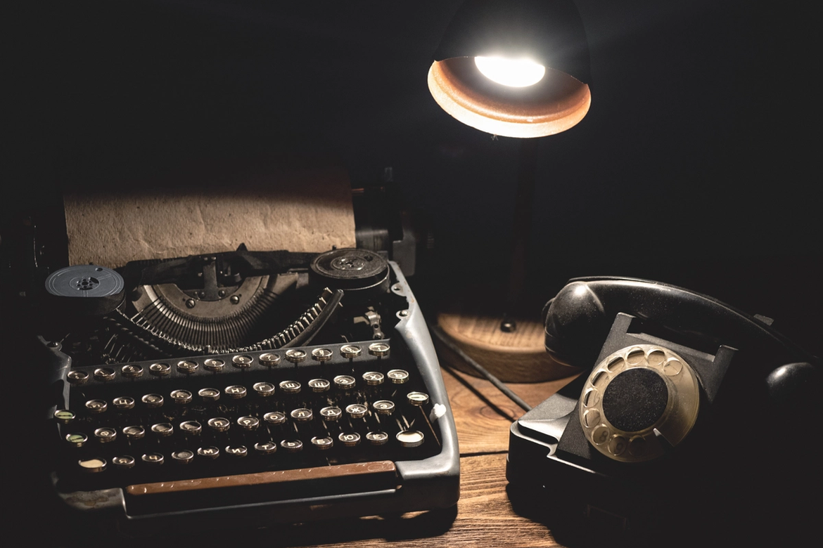 An old fashioned black mechanical typewriter with round keys, an old rotary black telephone and a black desk lamp lit up sitting on a desk in the dark.