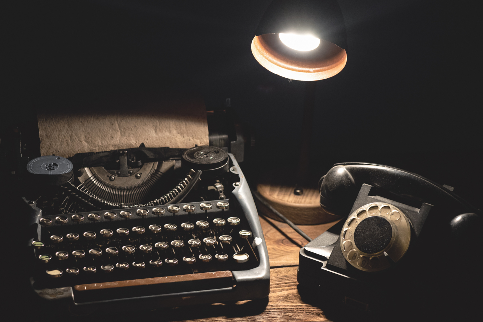 An old fashioned black mechanical typewriter with round keys, an old rotary black telephone and a black desk lamp lit up sitting on a desk in the dark.