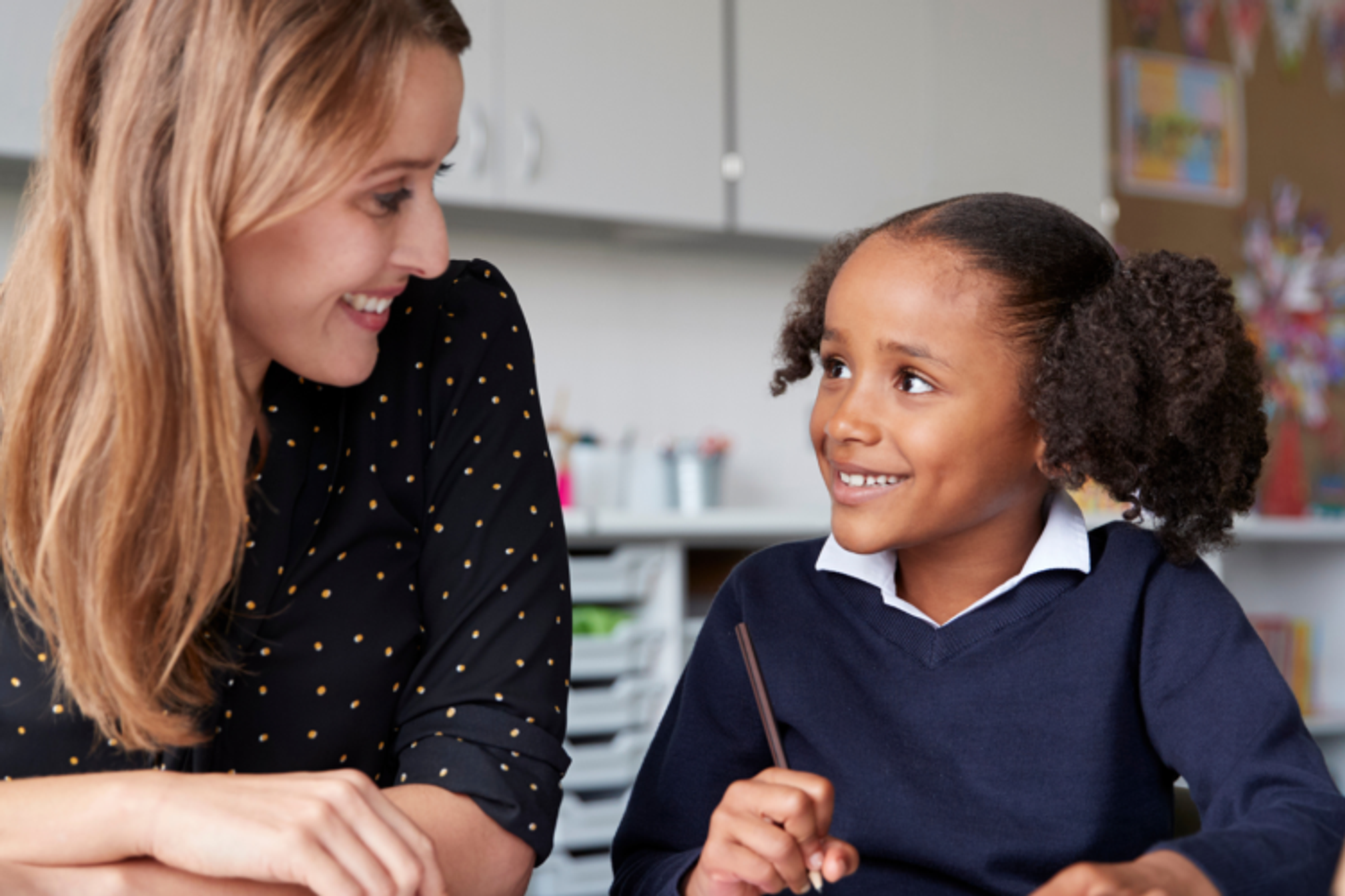 A woman is sitting with a young schoolgirl at a table, the girl is holding a pencil and they are looking at each other, smiling..