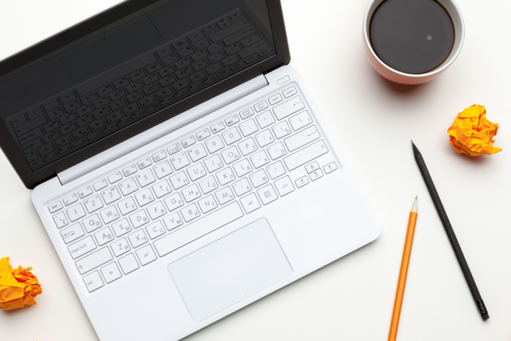 A laptop on a table with screwed up balls of paper, some pencils and a coffee cup.