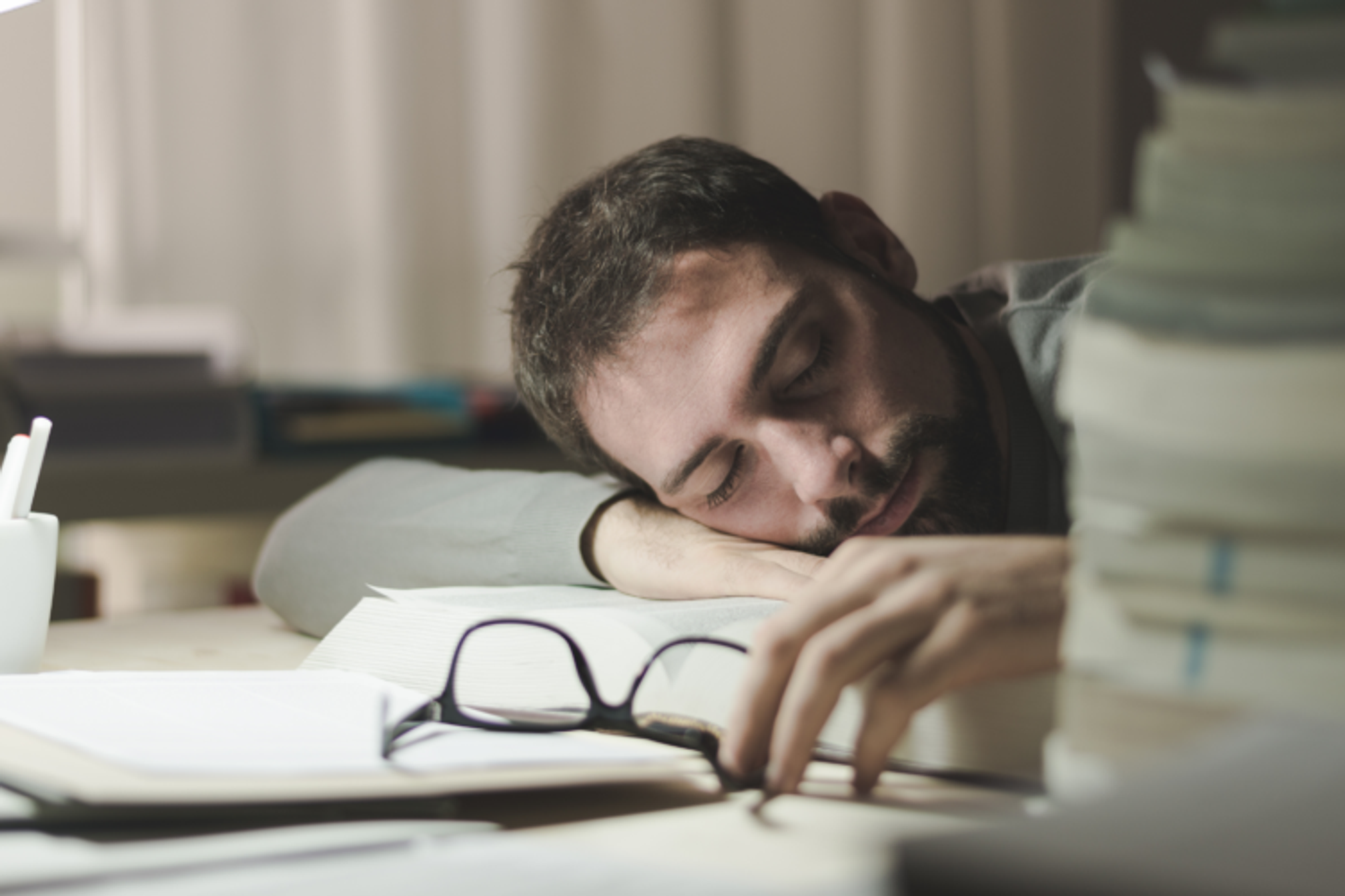 A man at a desk surrounded by books, he is asleep with his head on his work, he is holding his spectacles in his hand.