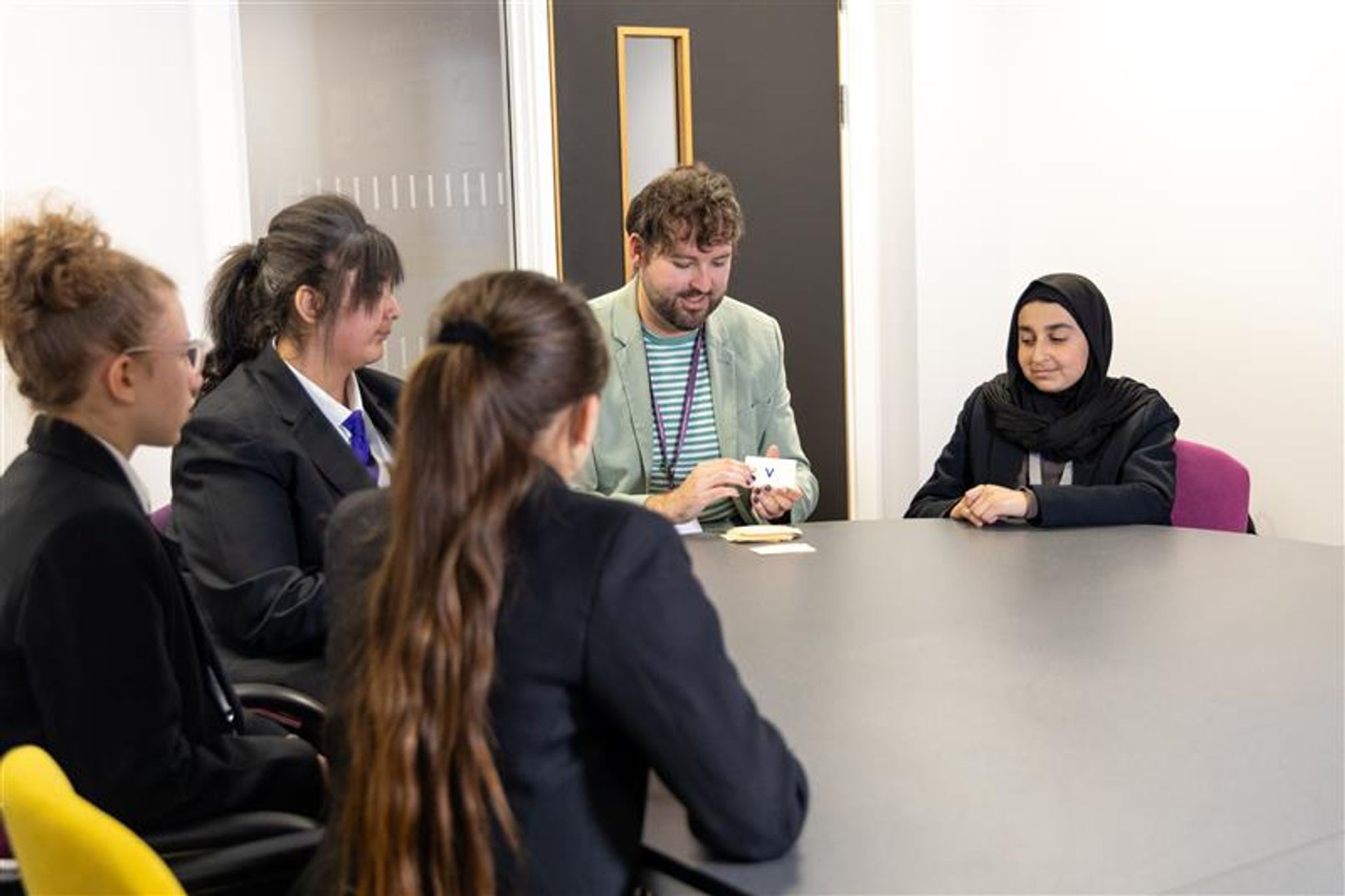 Secondary school children are sitting around a table in a classroom with a teacher, they are all looking at the teacher who is holding cards with letters on.
