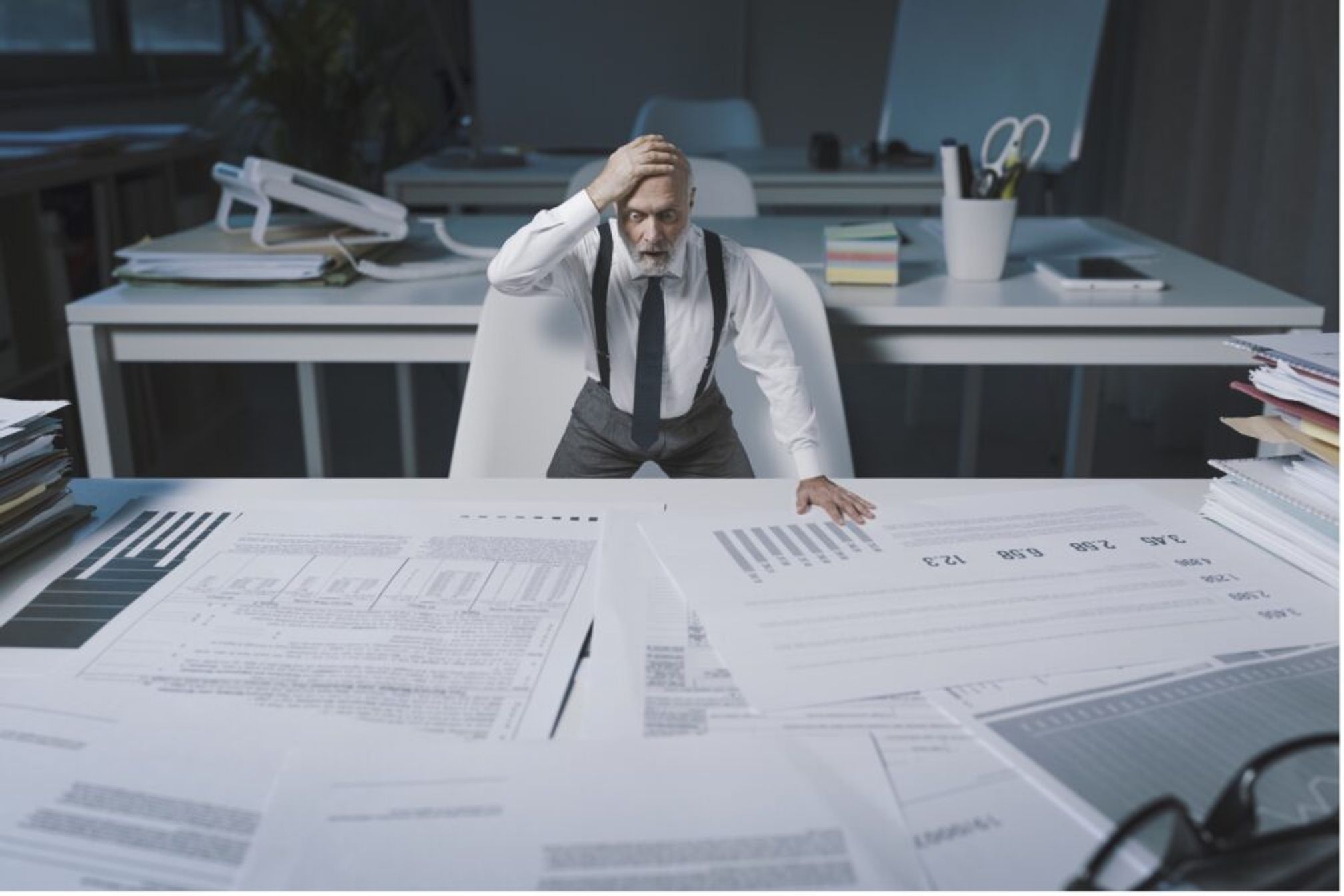 A businessman at a large table covered in pages of charts, reports and graphs, he has his hand on his head in despair.