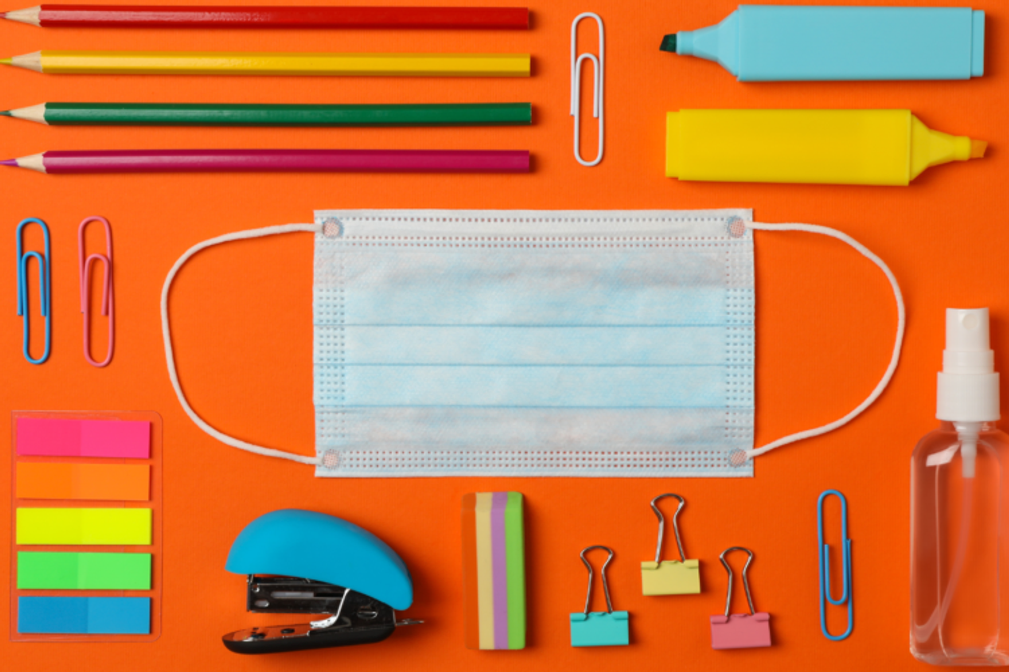 Colourful stationary: sticky notes, paperclips, stapler, pencils, eraser, bulldog clips and highlighter pens arranged neatly on a bright orange background alongside a clinical face mask and hand sanitiser spray.