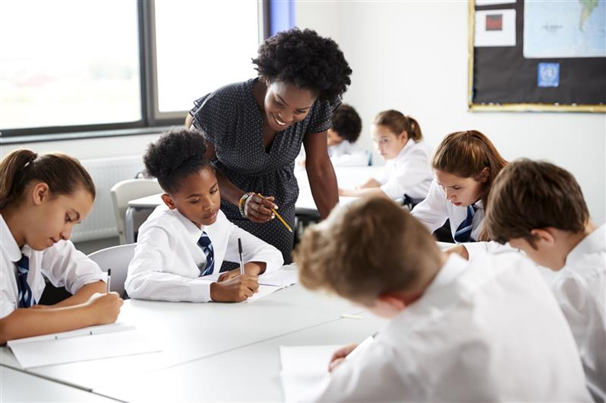 A classroom of young secondary school students in uniform writing on paper. A teacher is leaning over to talk to one student and is looking at her work.