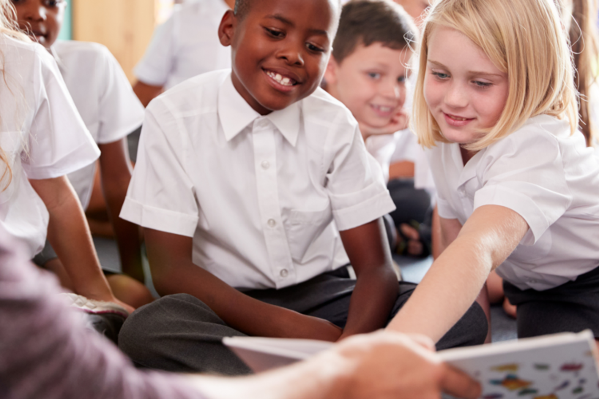 Primary school children in class looking at a book that a teacher is holding, one girl is pointing to something in the book.