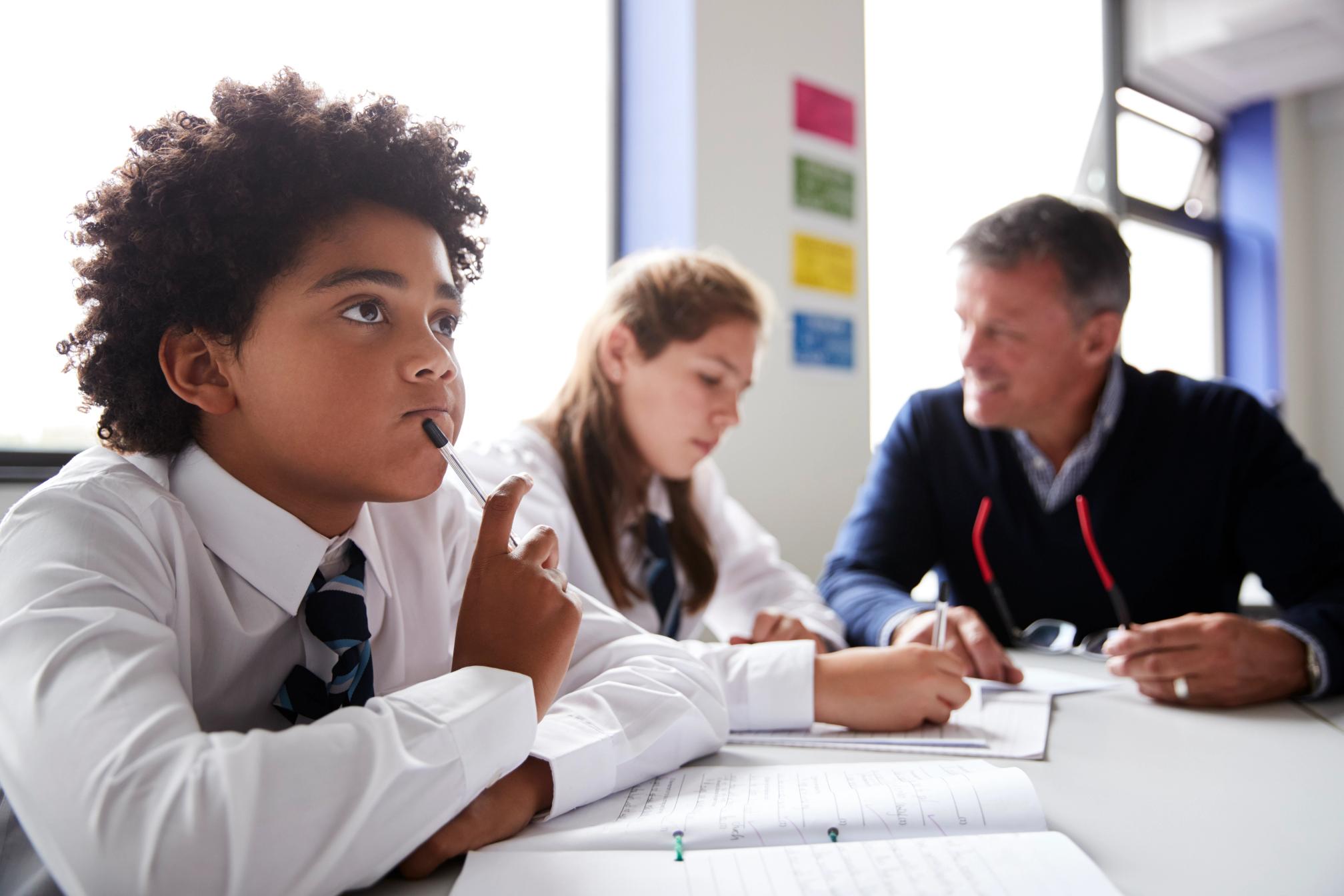 School children sitting at a desk writing, one is speaking to a teacher