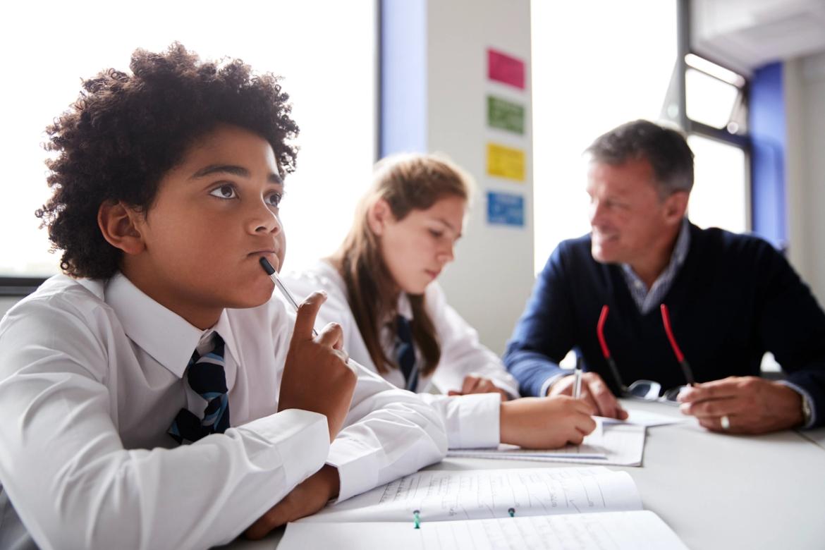 School children sitting at a desk writing, one is speaking to a teacher