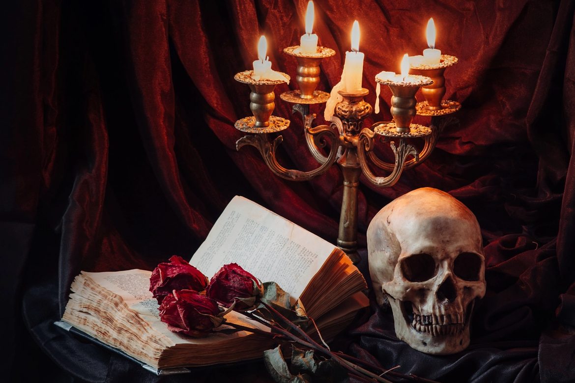 A human skull, an old book with some dying red roses placed on it and a candelabra with 5 lit candles displayed on a black velvet cloth with a red velvet drape behind.
