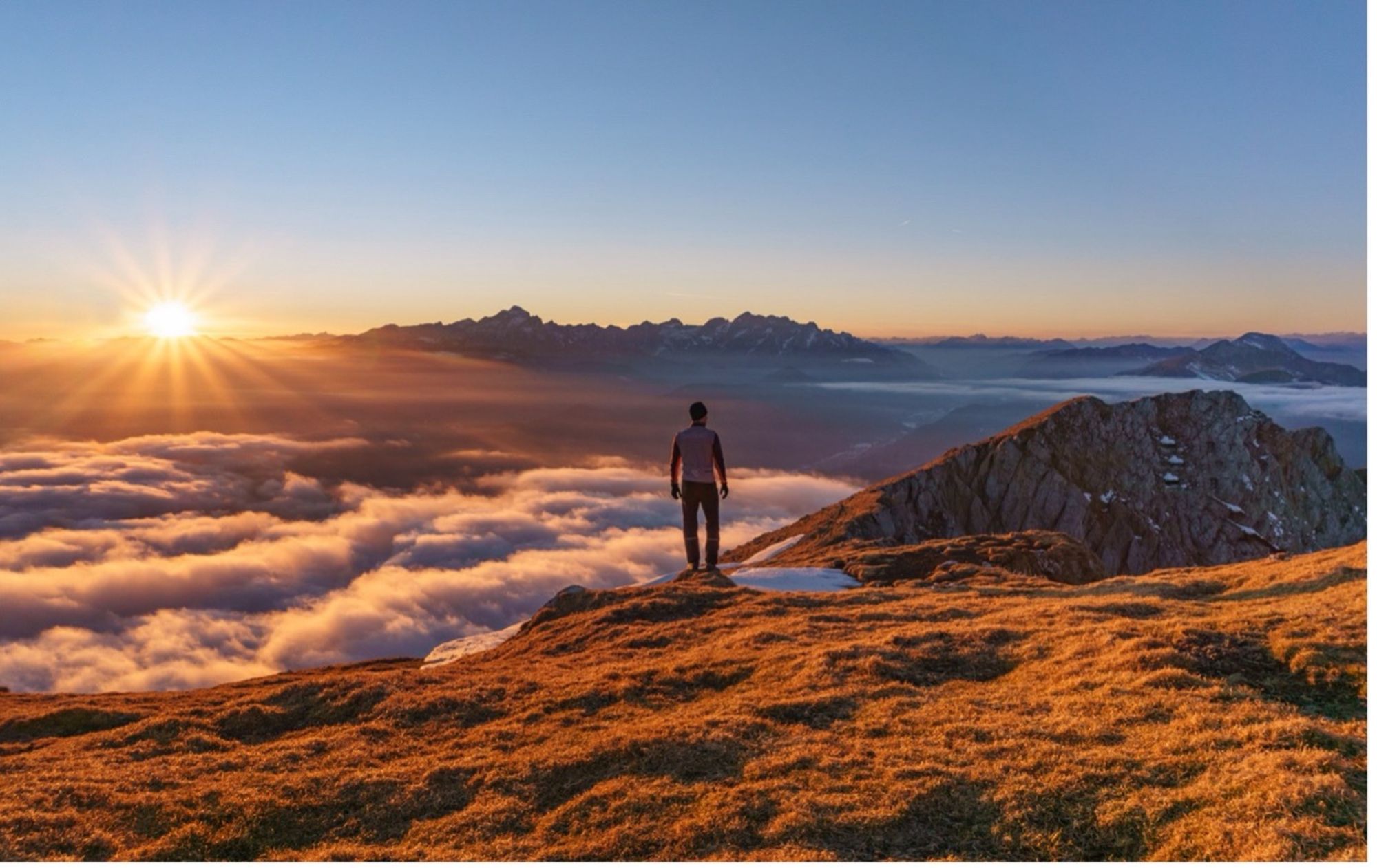 A man standing on the edge of a high mountain looking at the sunrise with the clouds beneath him.