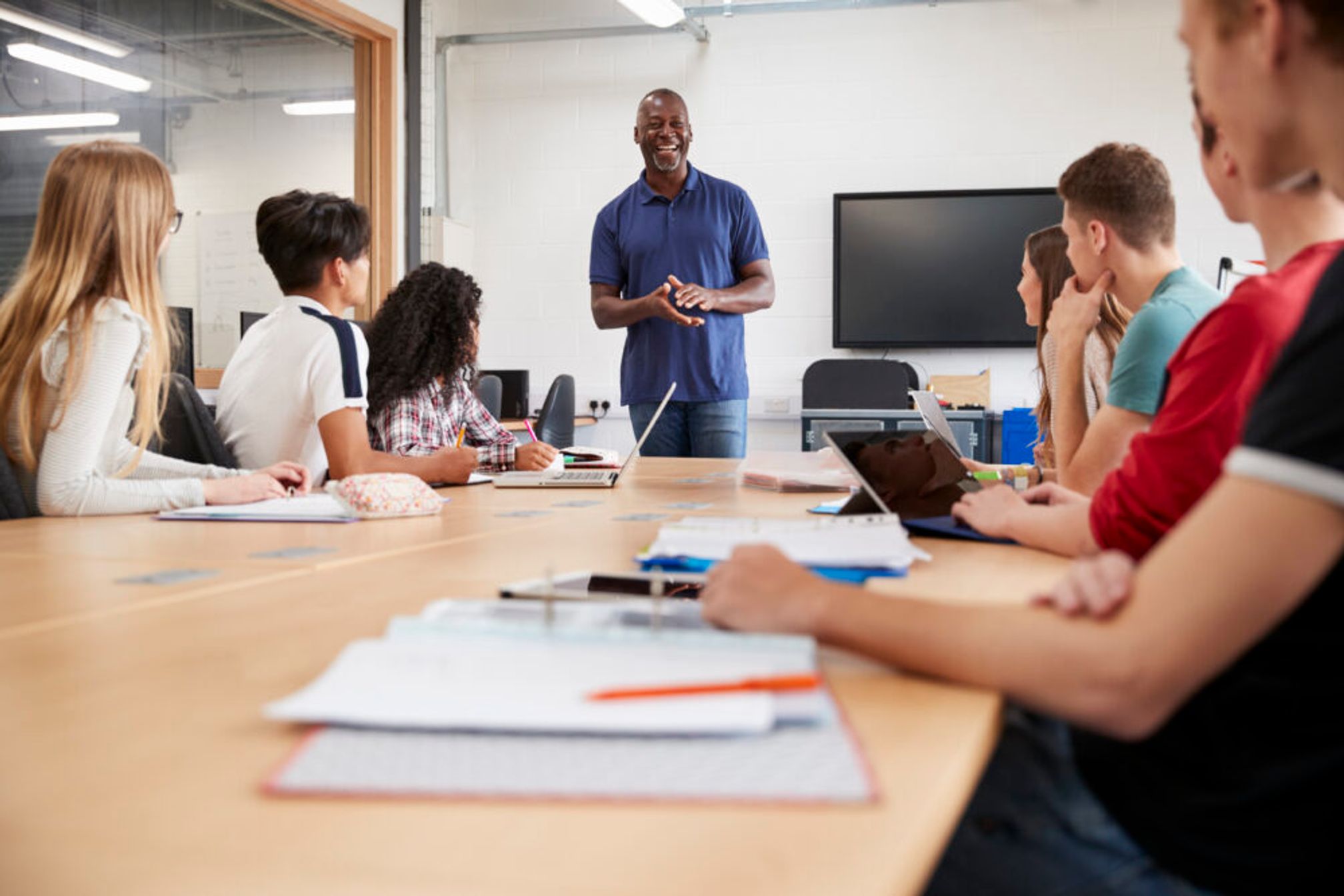 College students sitting around a long table in a classroom looking at their teacher who is speaking to them and smiling.